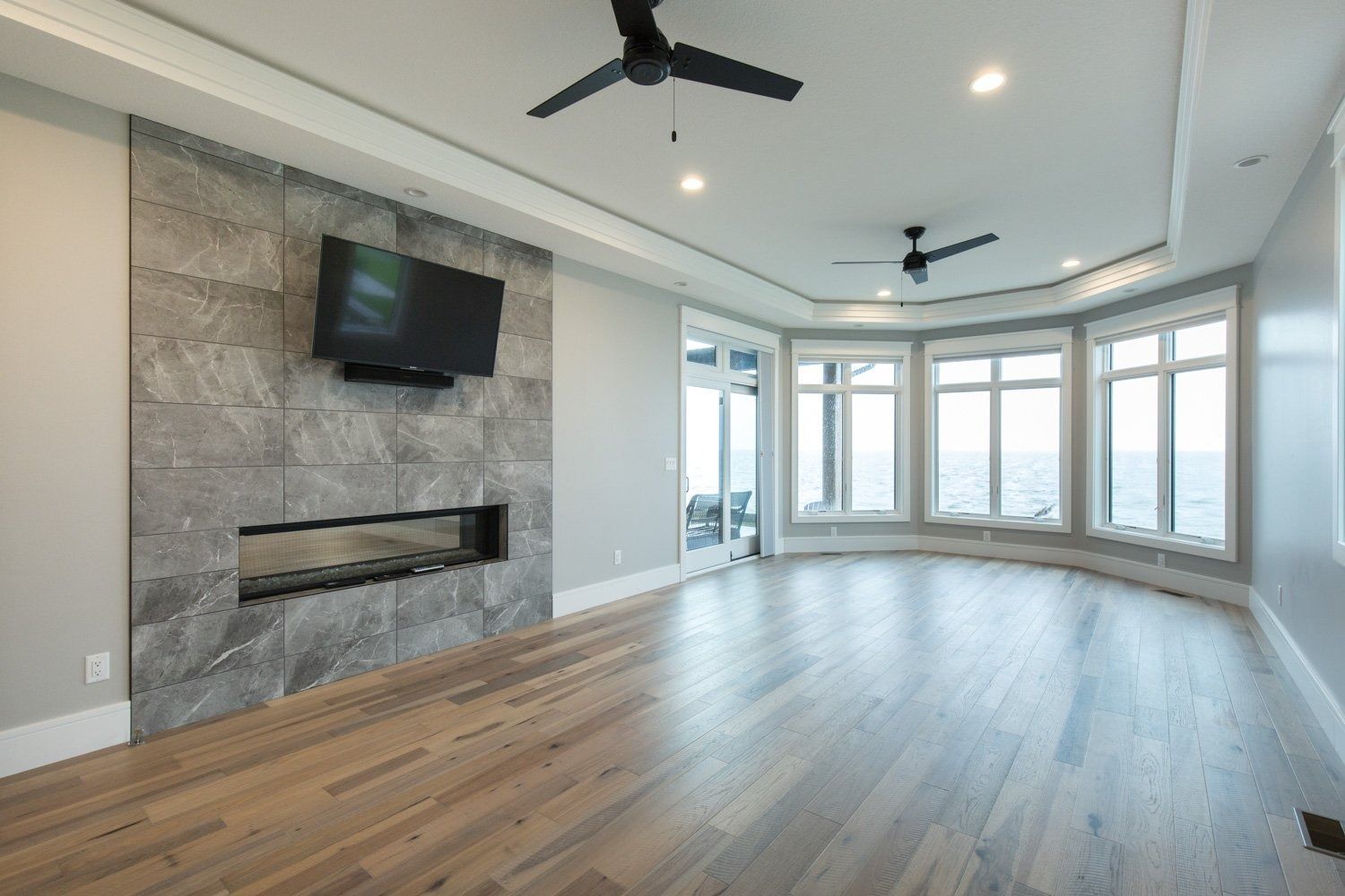 Empty living room with wood floors, gray fireplace wall, and large windows.