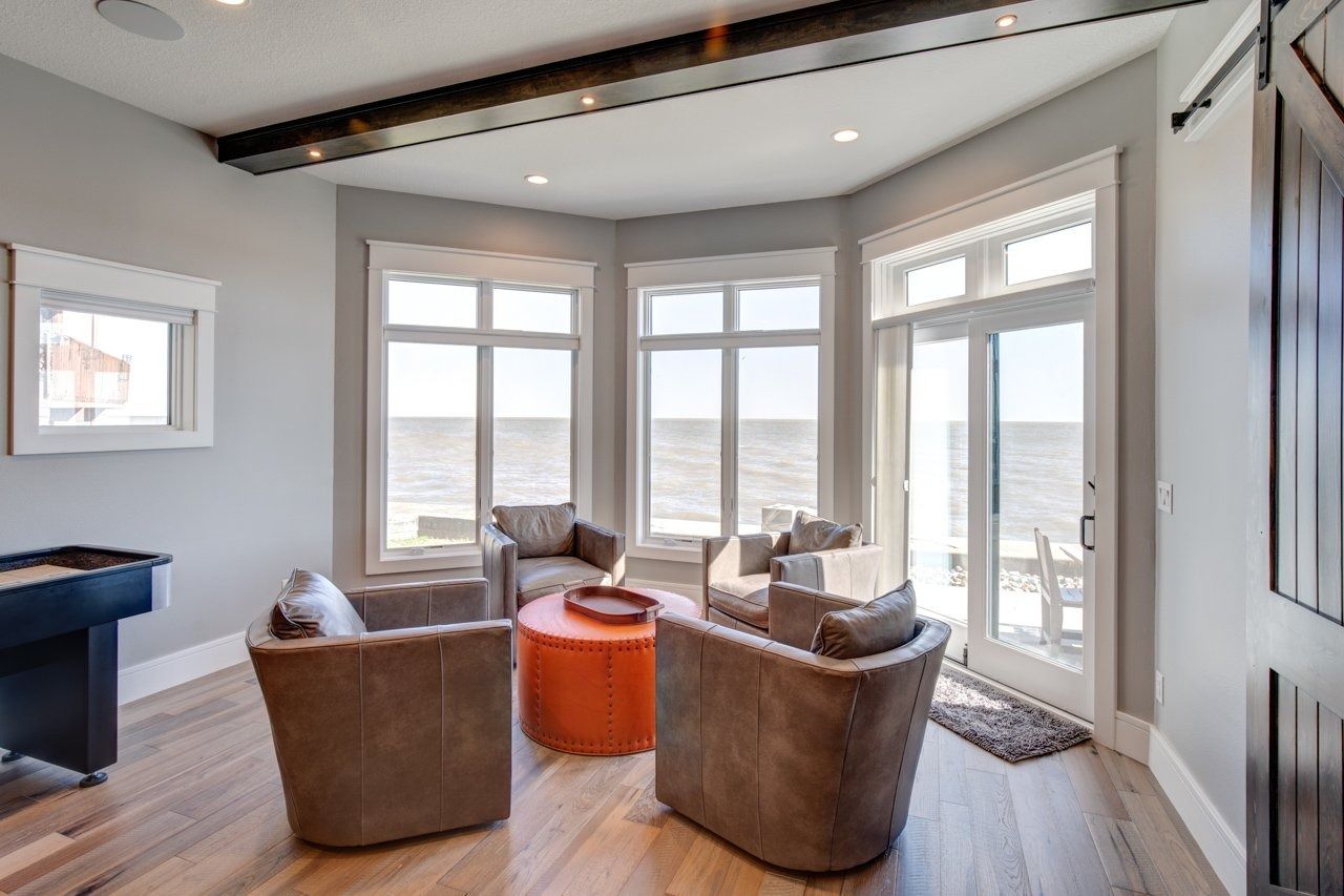 A sunroom with a waterfront view. Gray walls, wood floor, four brown leather chairs, and an orange coffee table.