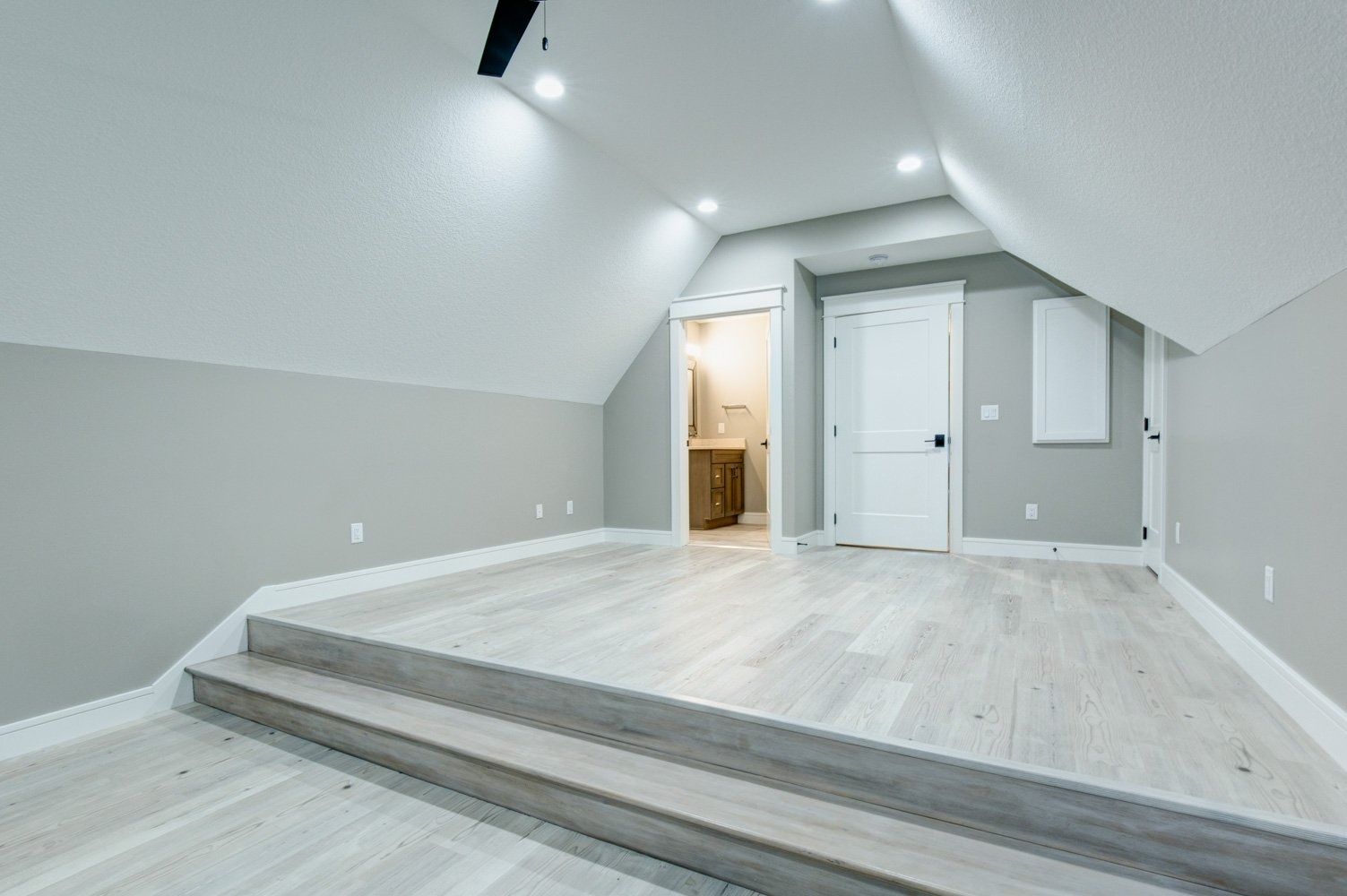 Empty, modern room with sloped ceiling, light wood floor, and recessed lighting. A bathroom doorway is visible.