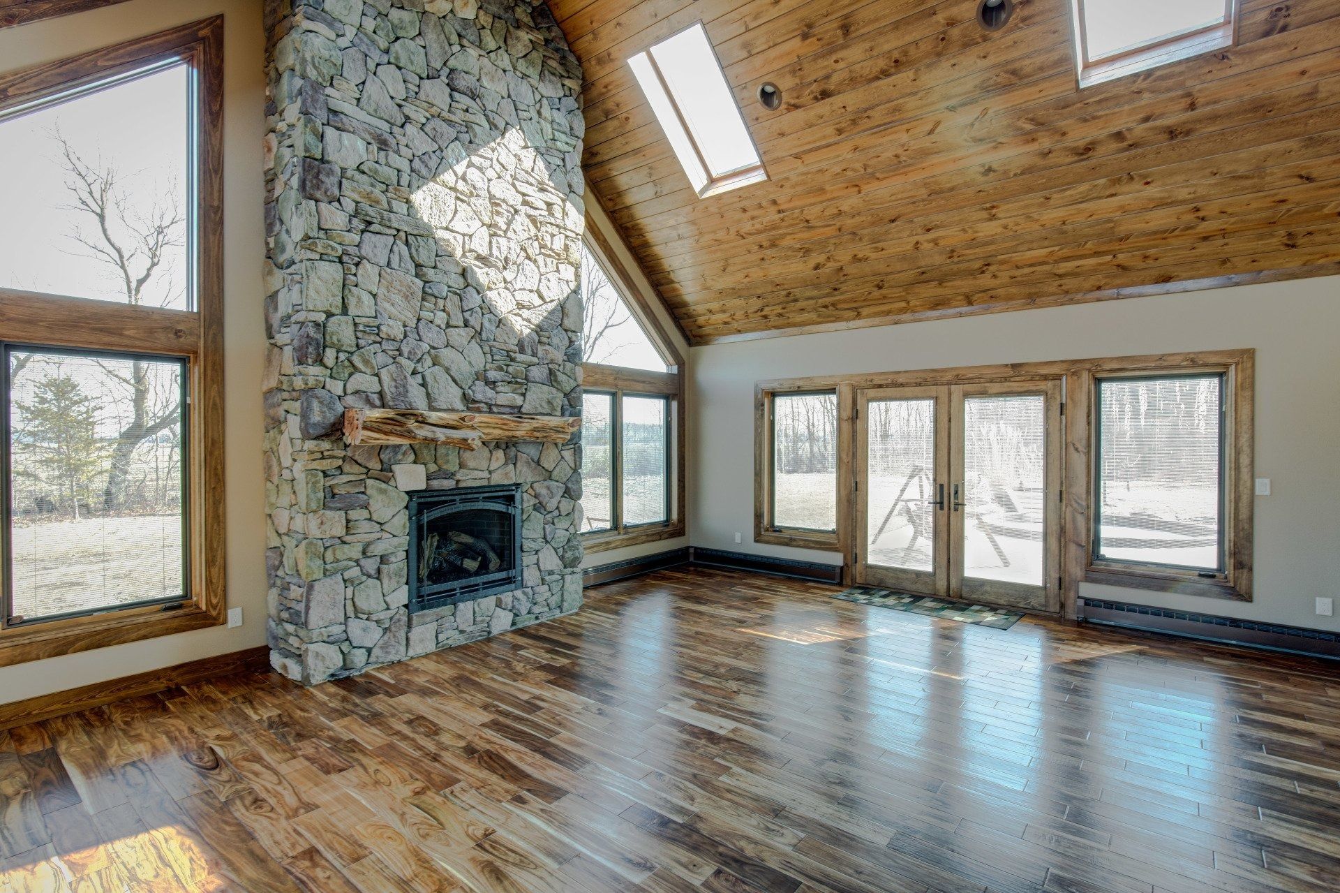 Spacious room with stone fireplace, wood floors, large windows, and a vaulted, wood-paneled ceiling.