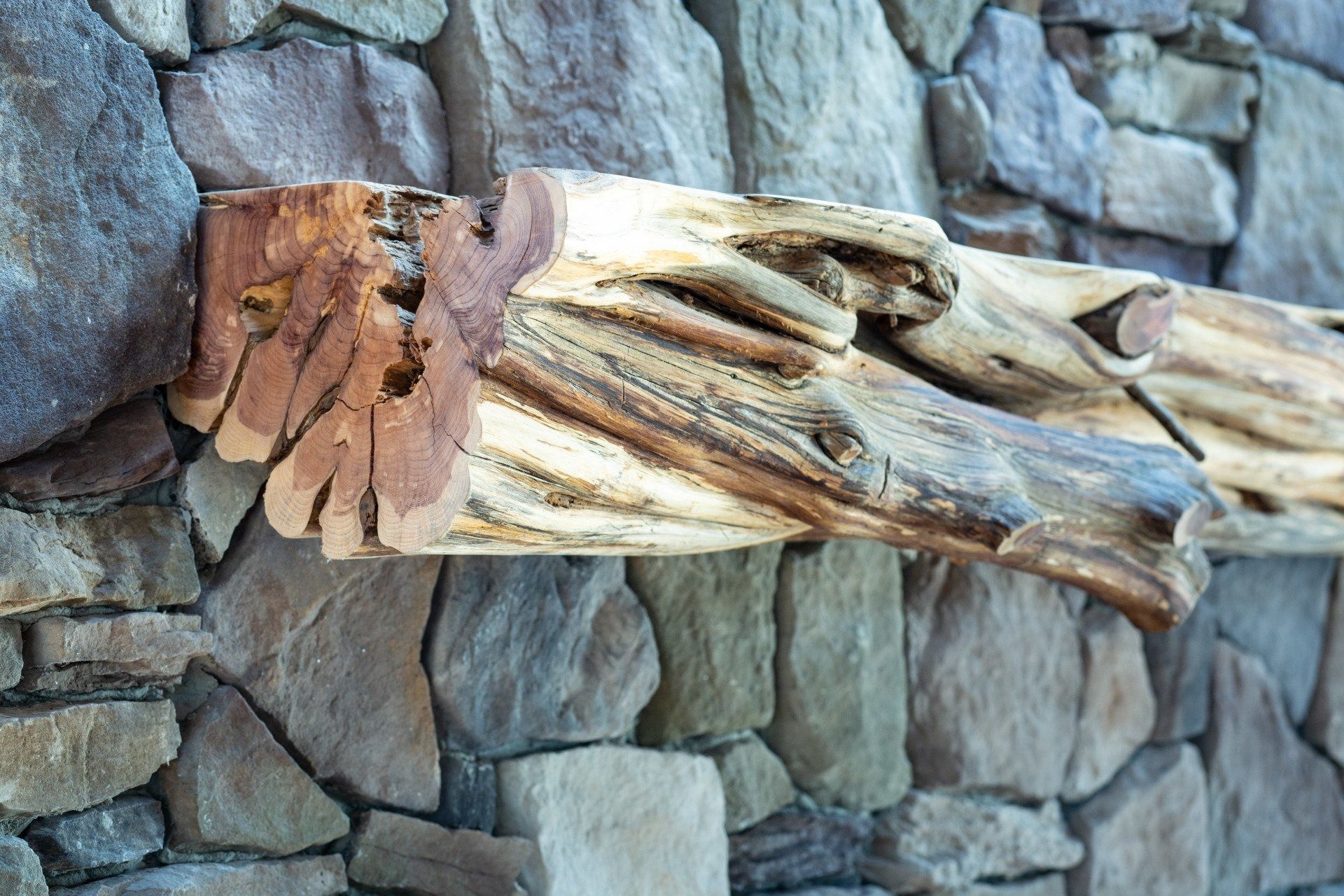 Rough-hewn wooden beam mantel on a stone wall. The wood is light and dark brown.