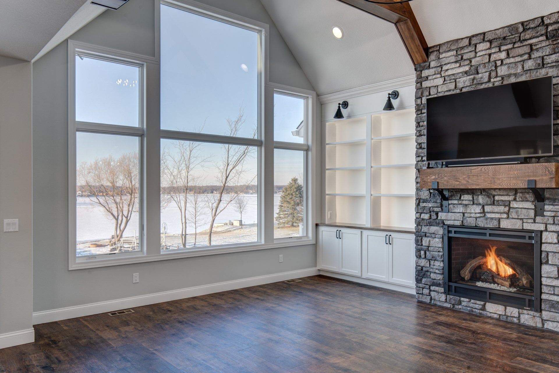 Living room with large windows overlooking a lake, fireplace, built-in shelves, and dark wood floors.