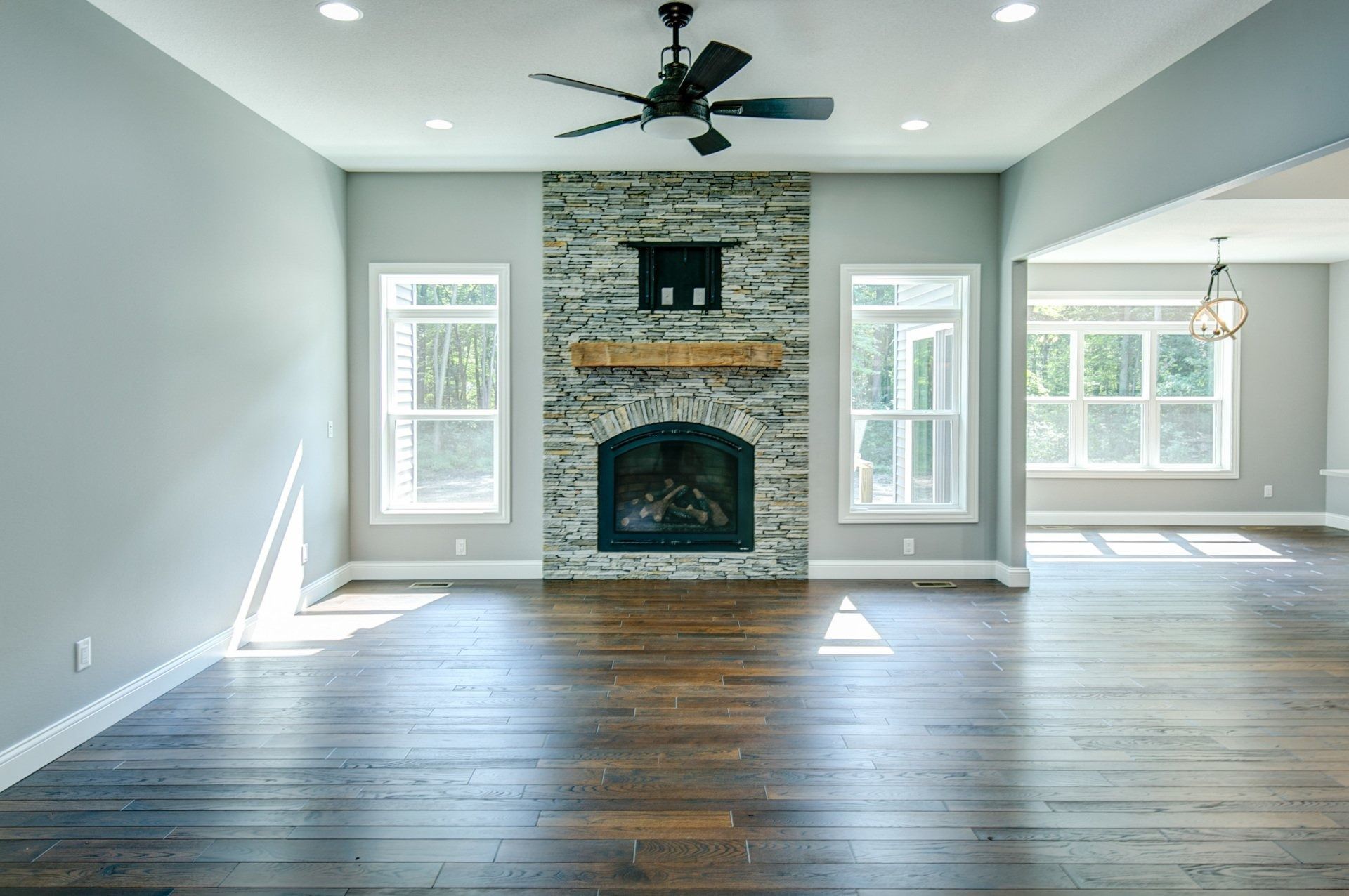Empty living room with fireplace, windows, ceiling fan, and hardwood floors; light gray walls.