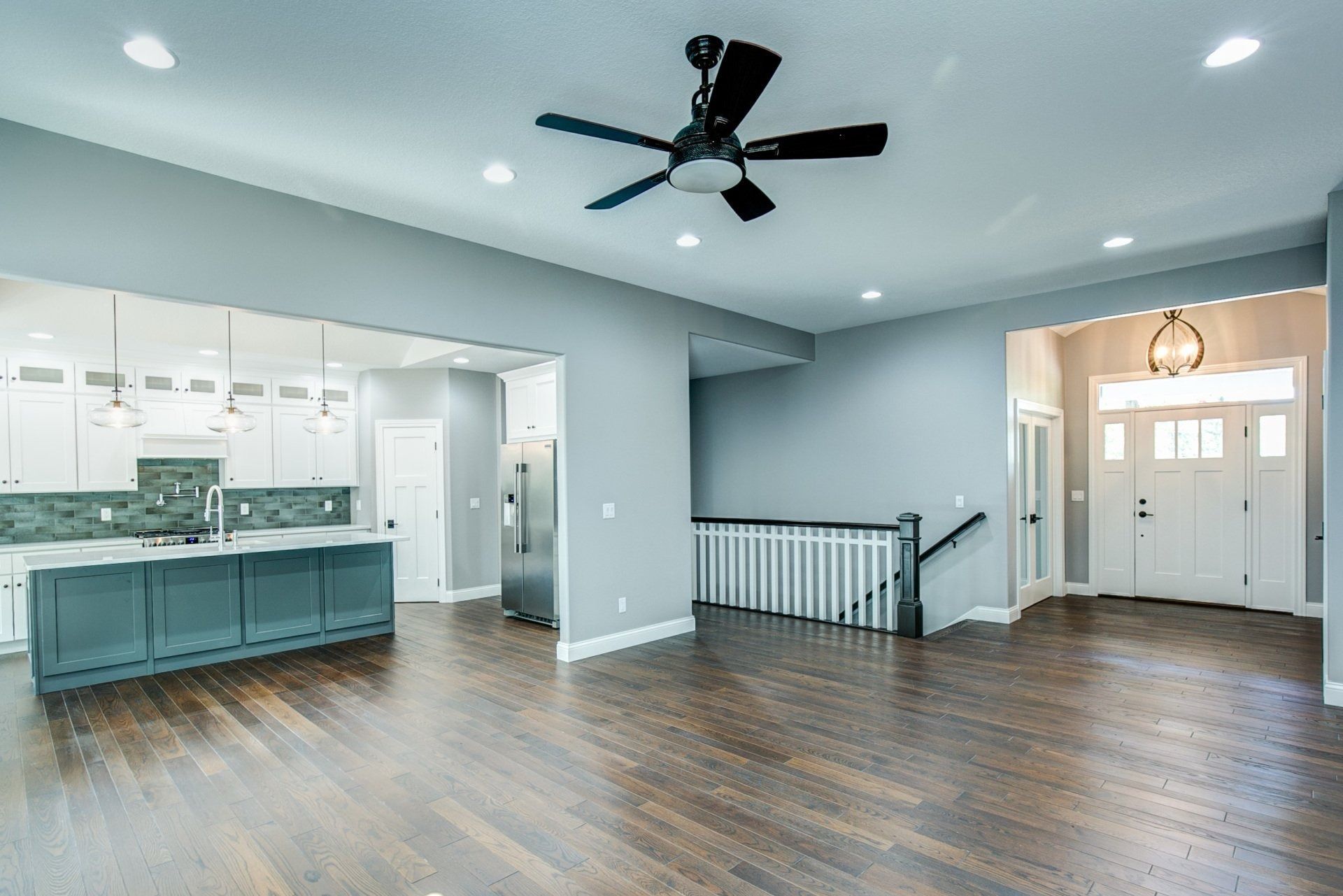 Spacious living area with dark wood floors, open to kitchen and entrance. Gray walls, ceiling fan, and white trim.