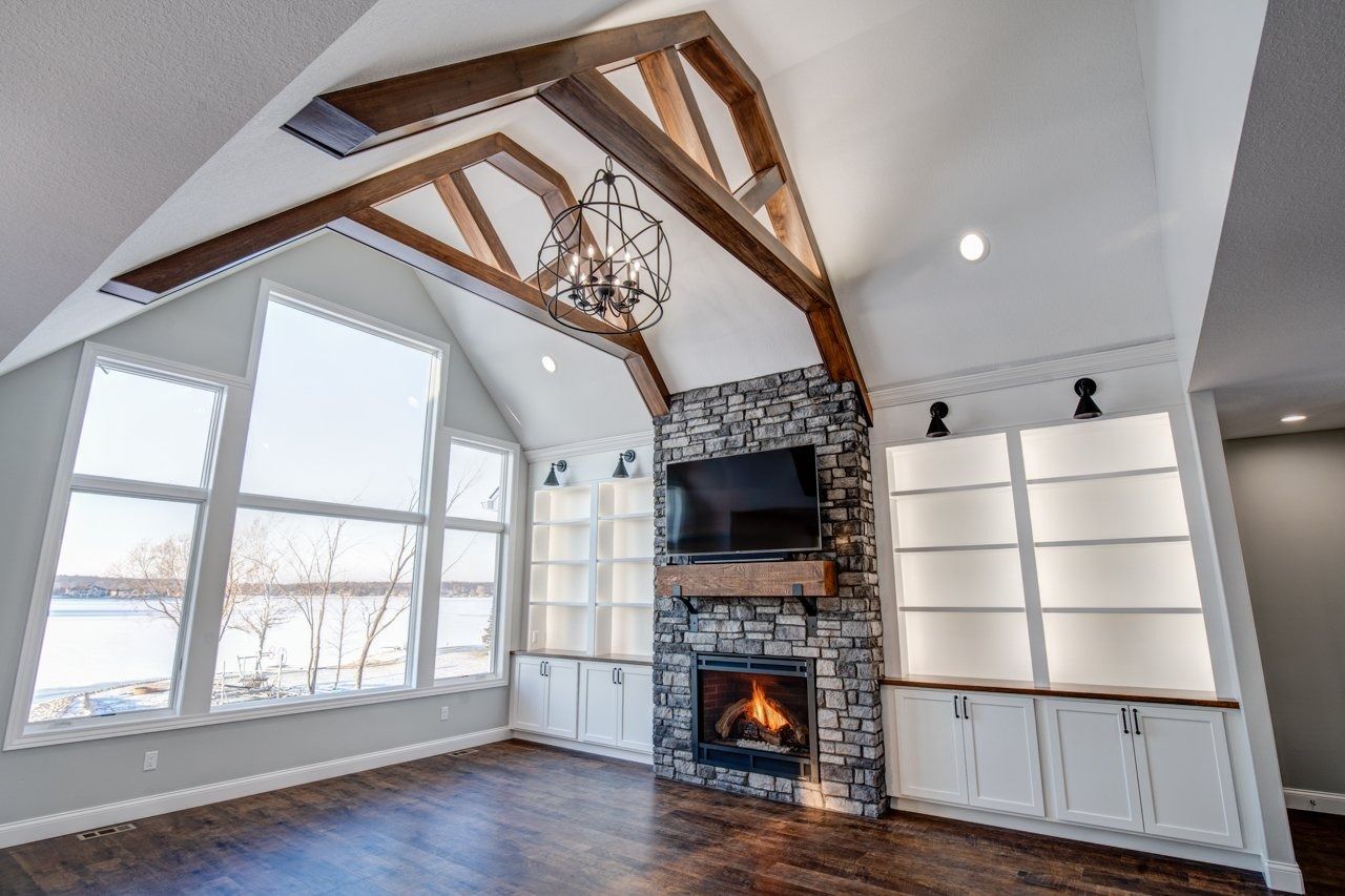 Living room with a stone fireplace, built-in shelves, large windows, and exposed wooden beams.