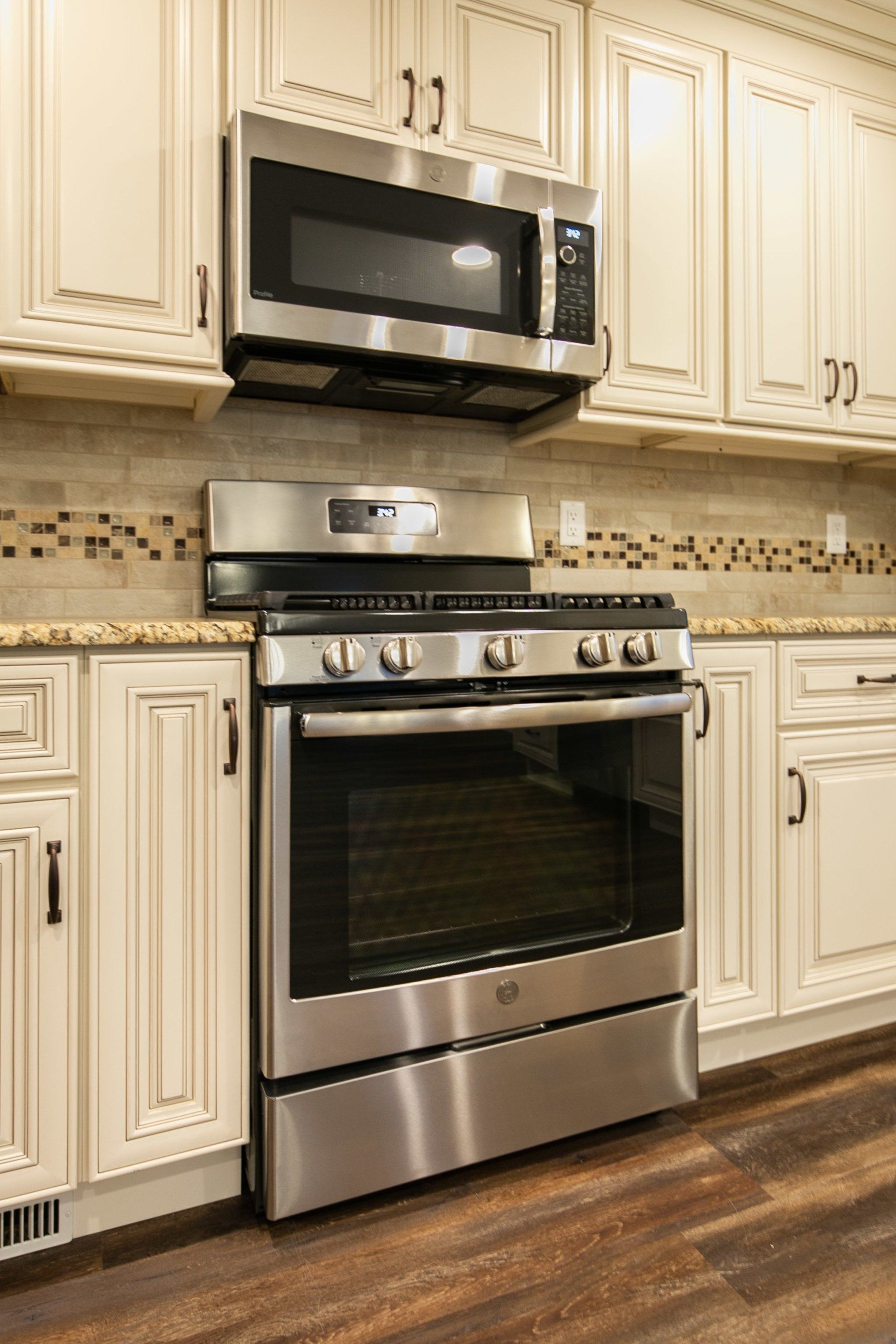 Stainless steel range and microwave beneath cream-colored kitchen cabinets with dark hardware and a tiled backsplash.