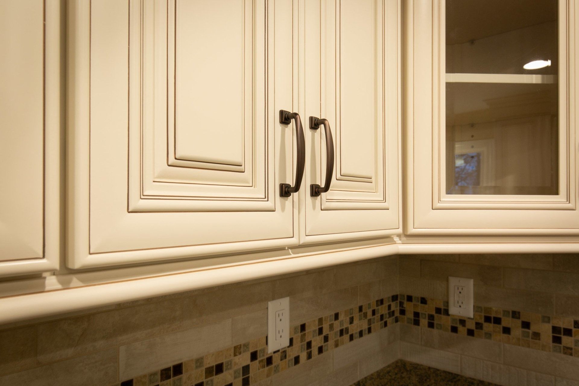 Cream-colored kitchen cabinets with dark handles above a countertop with mosaic backsplash.
