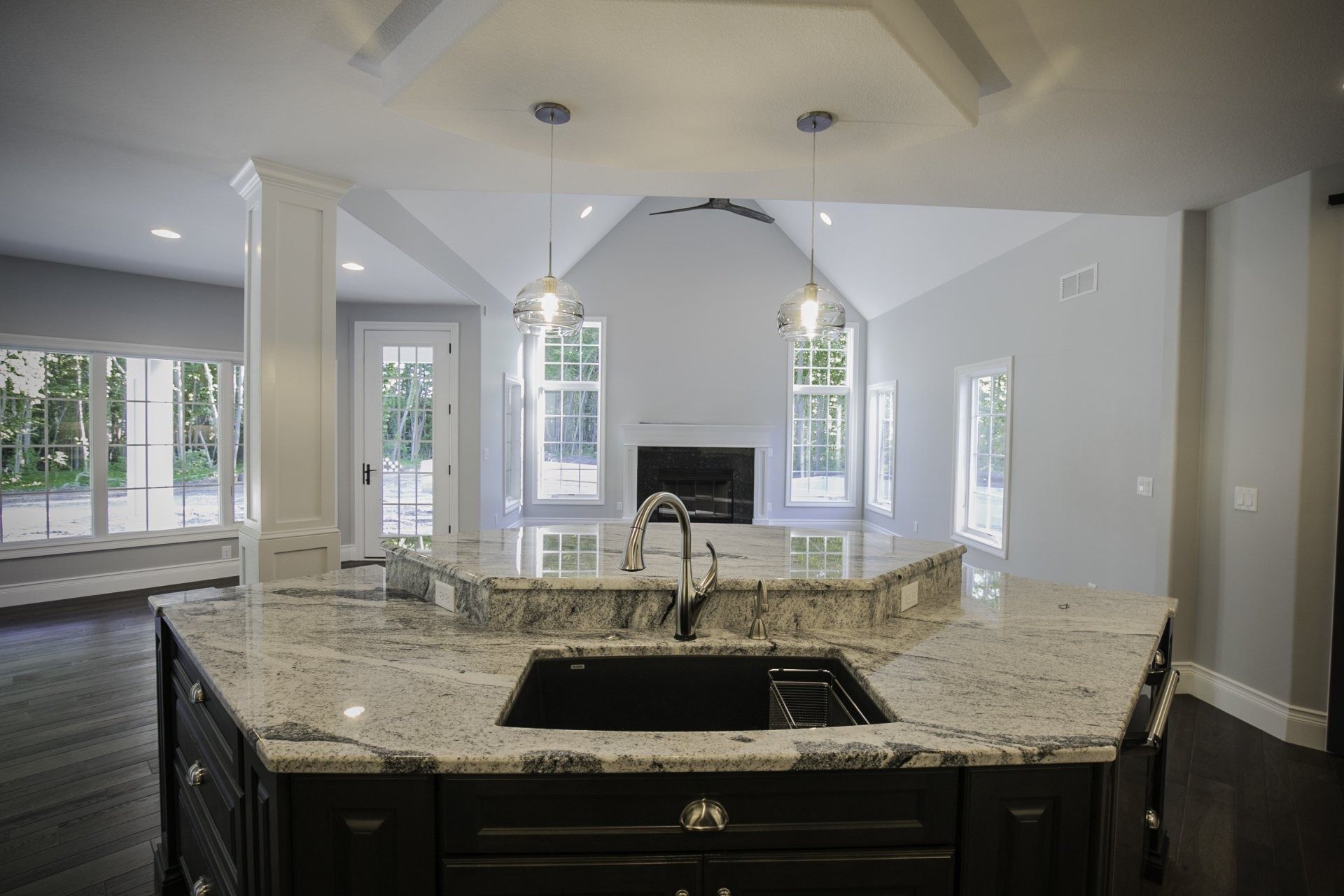 Kitchen with granite countertop island, dark cabinets, pendant lights, and living area in the background with a fireplace.