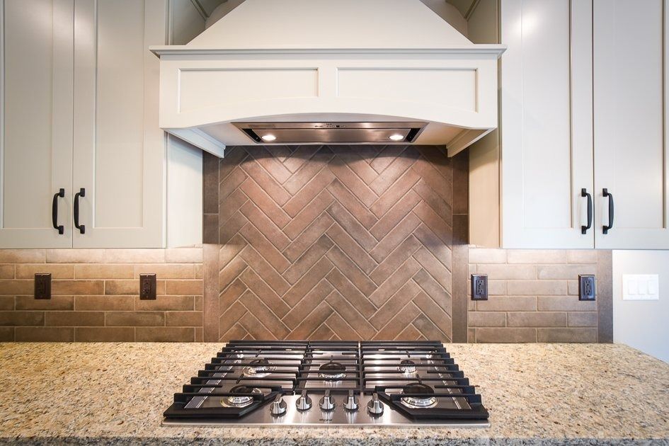 Kitchen with stove, gray cabinets, beige granite countertop, and brown herringbone tile backsplash.