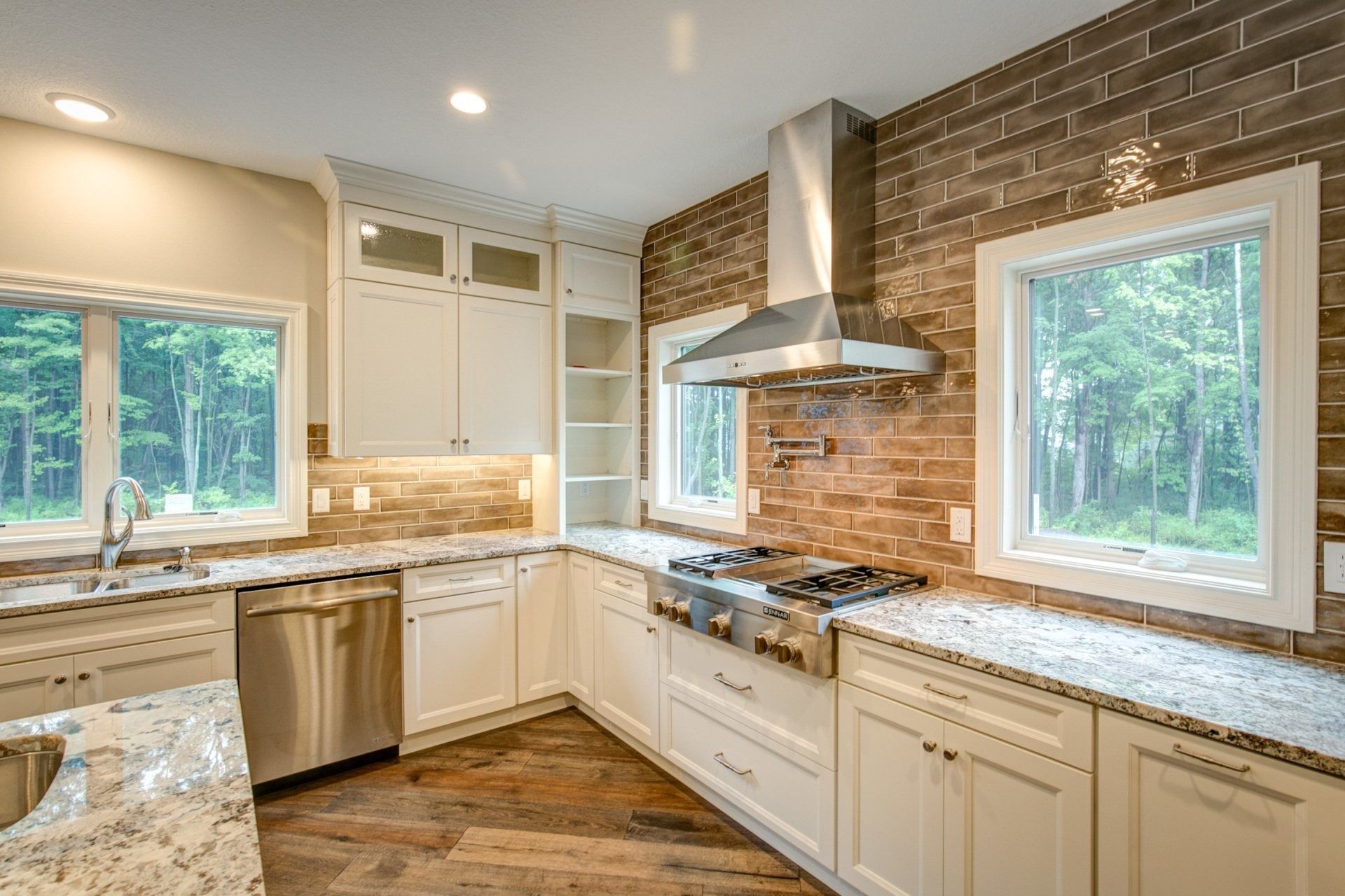 Modern kitchen with white cabinets, stainless steel appliances, brick backsplash, and two windows overlooking trees.