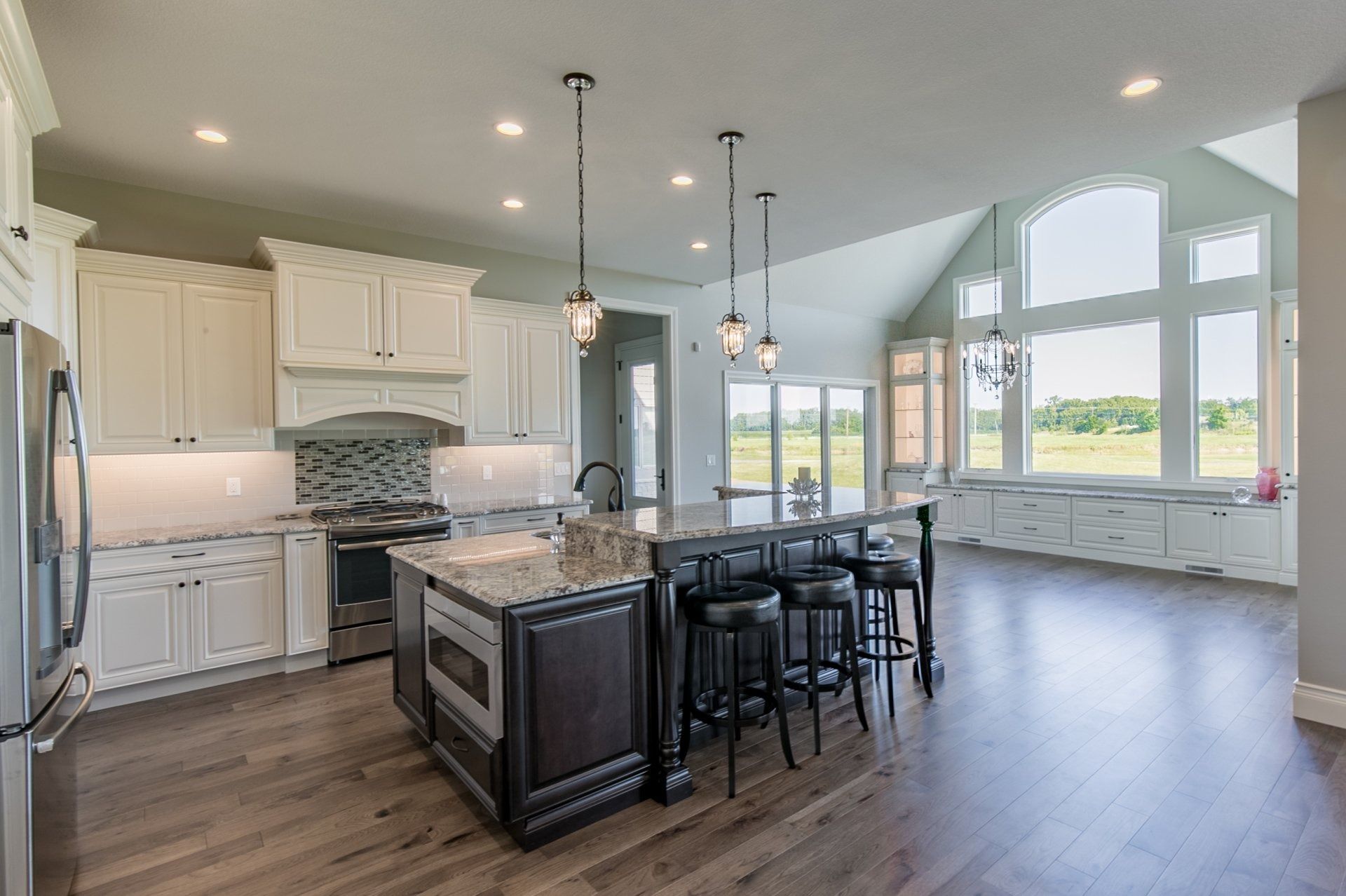 Spacious modern kitchen with white cabinets, dark island with seating, and large windows overlooking a field.