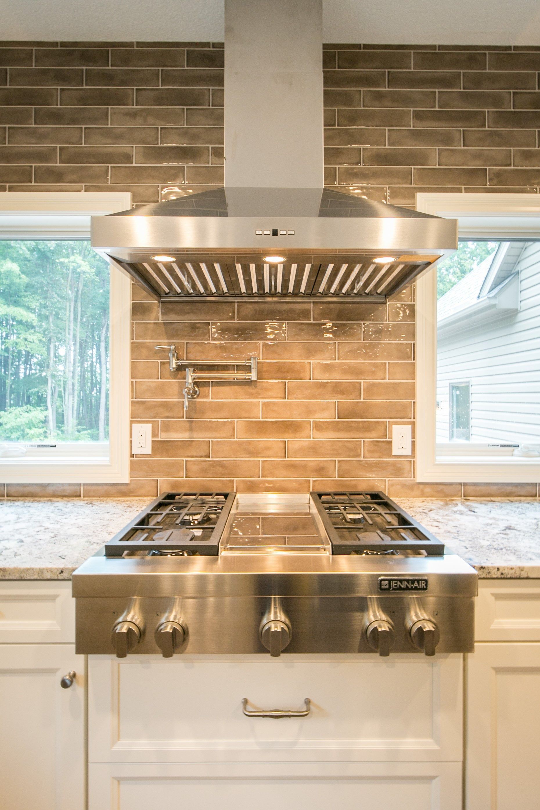 Stainless steel range and hood in a kitchen with brick backsplash and windows.