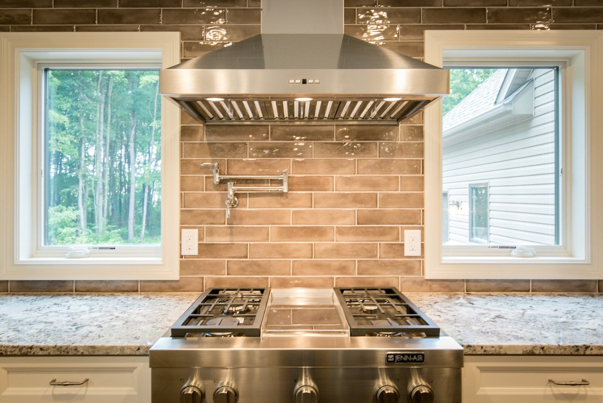 Stainless steel range and hood in a kitchen, flanked by windows with a brick-style backsplash.