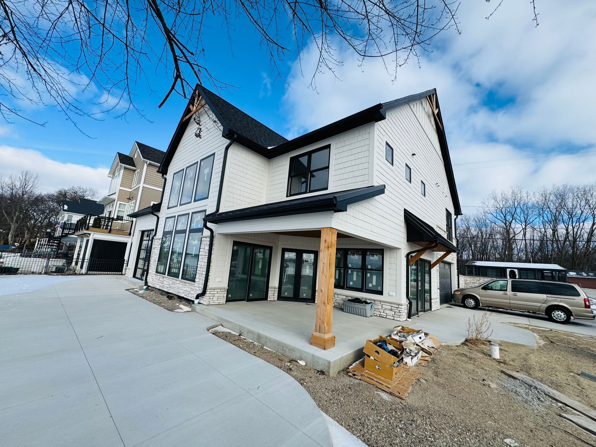White house with black trim, under construction. Gray concrete patio, blue sky, and a van.
