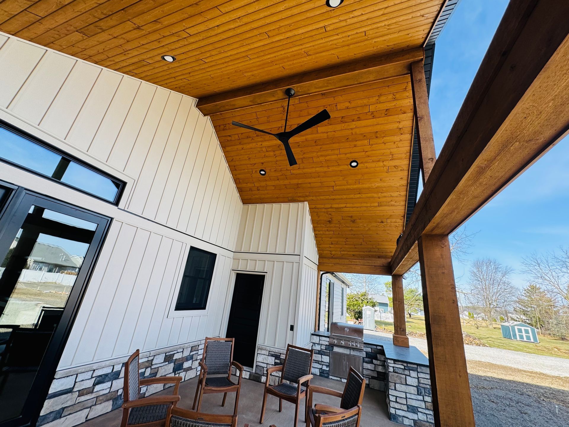 Covered porch with wood ceiling and black ceiling fan. White siding and stone accents. Several chairs are visible.