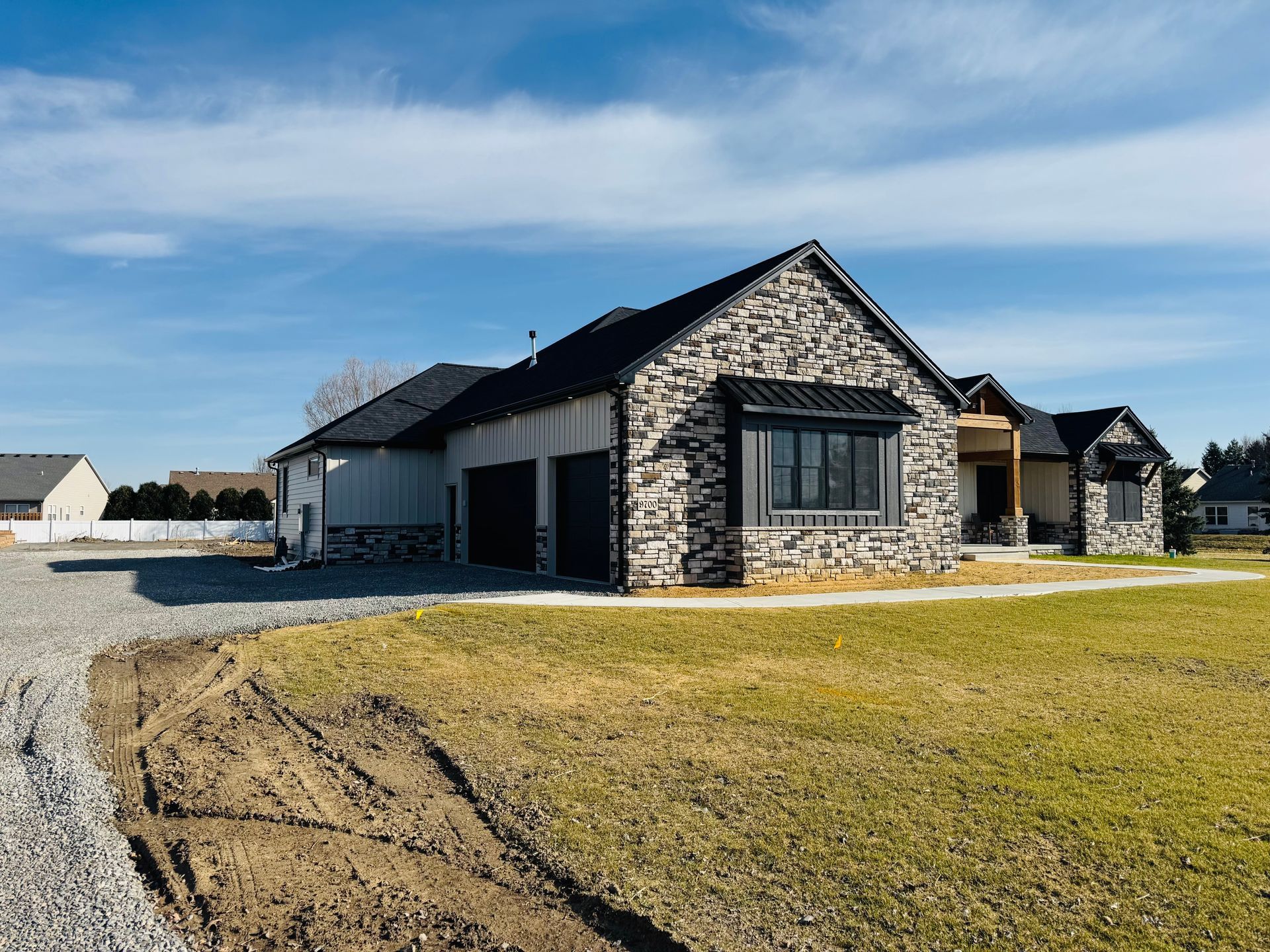 Stone-faced house with black roof and garage doors, sitting on a grassy lot, gravel driveway, under a blue sky.