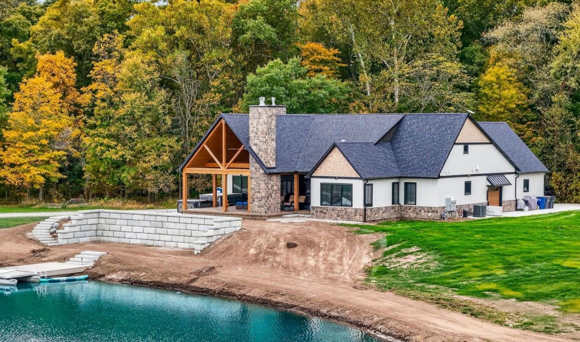 Modern white house with stone chimney and timber-framed porch by a pond, surrounded by fall foliage.