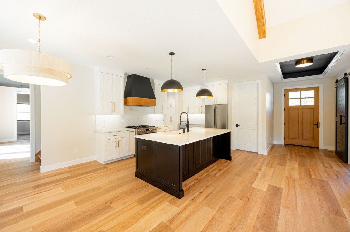 Modern kitchen with island, white cabinets, black range hood, wood floors, and pendant lights.