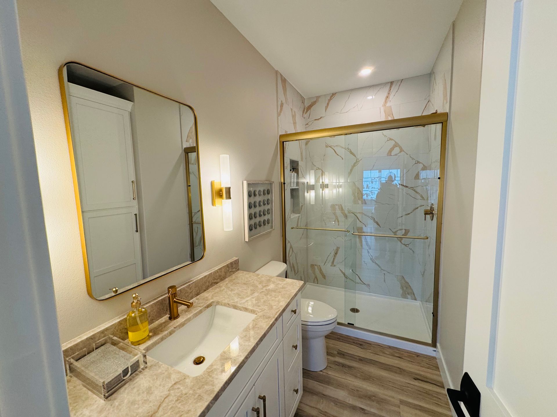 Bathroom with gold fixtures, marble-look shower and countertop, white vanity, and a large mirror.