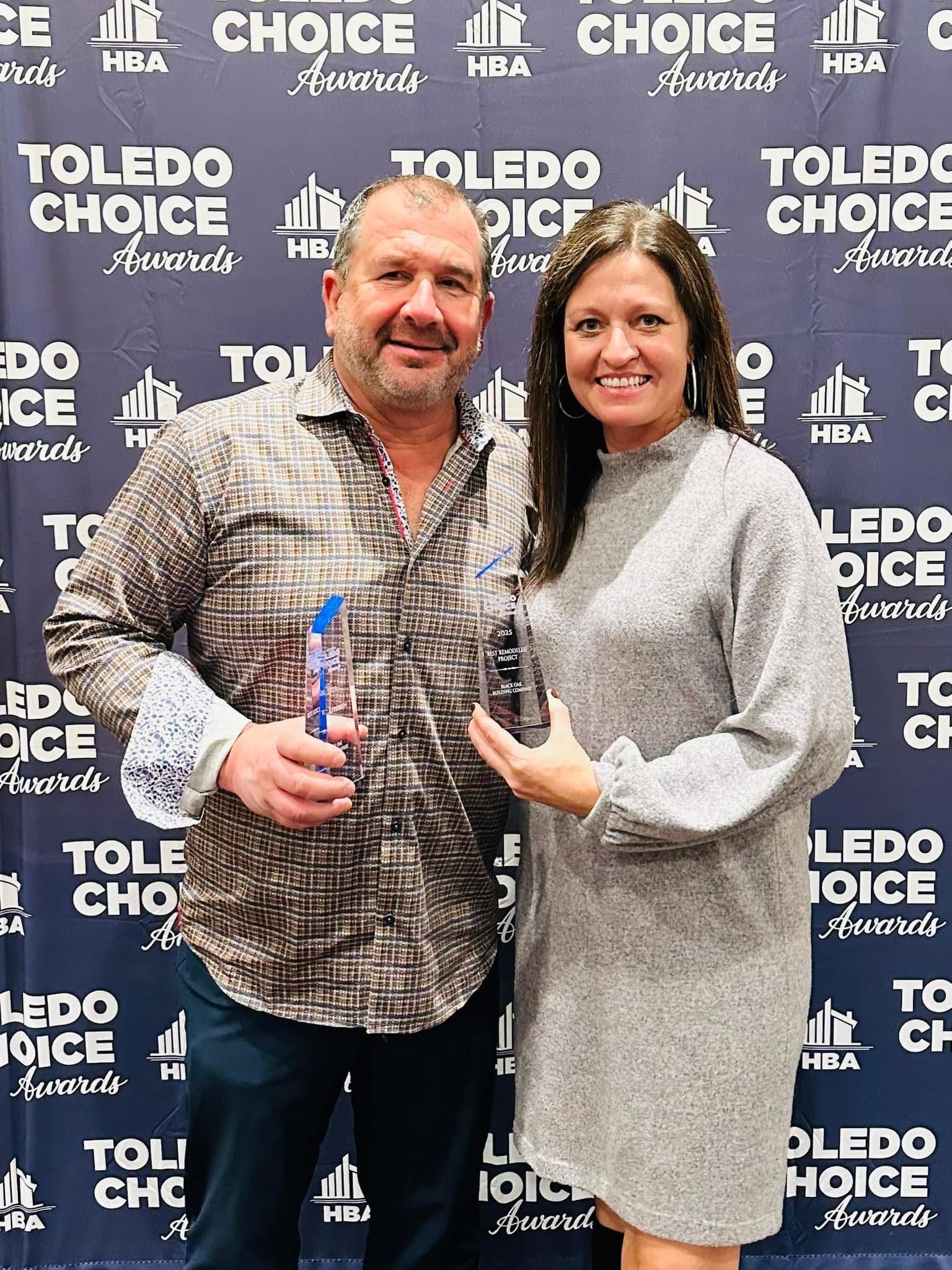 A man and woman smiling while holding glass award trophies in front of a blue 