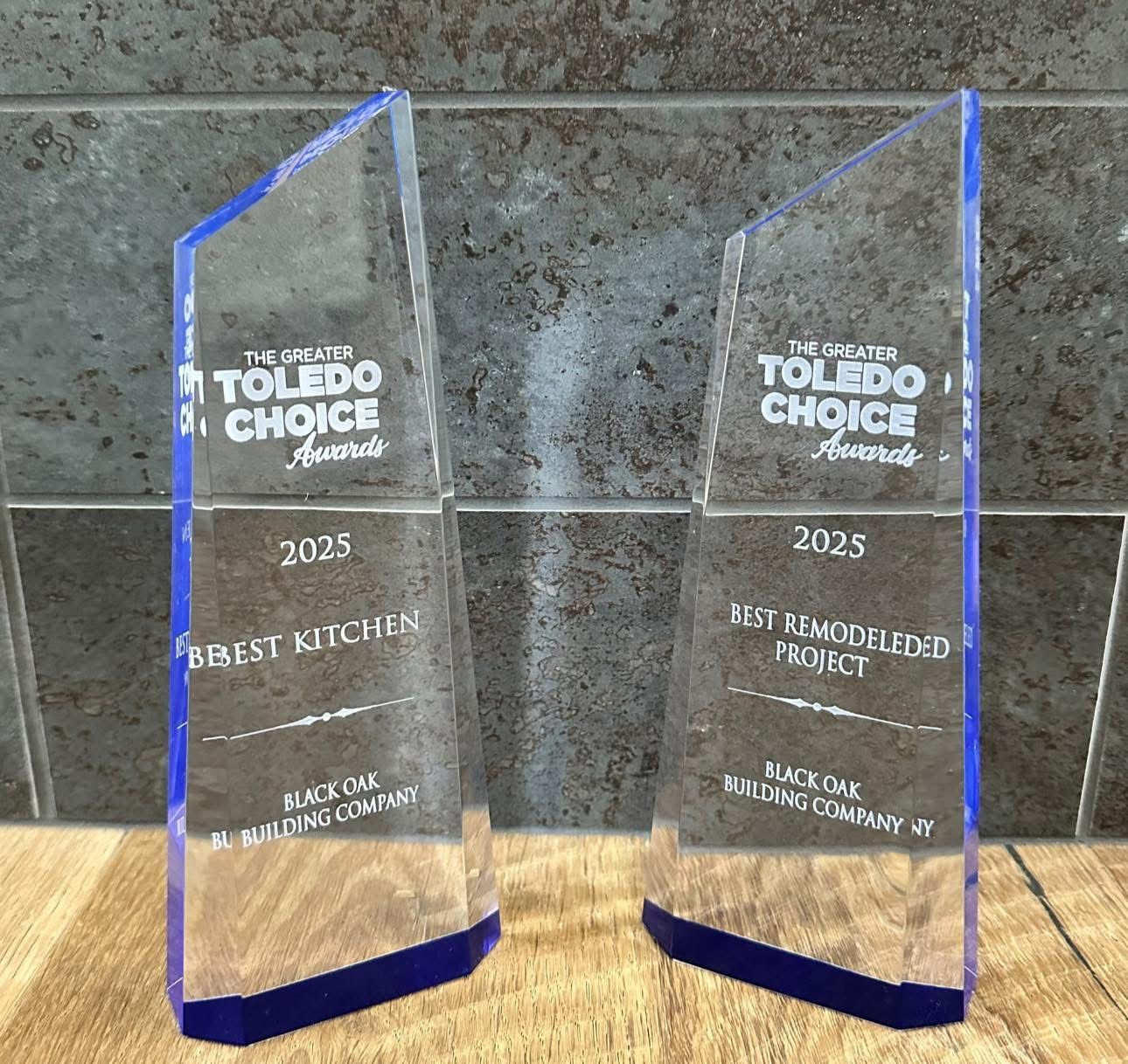 Two clear, blue-tinted 2025 Toledo Choice Awards trophies on a wooden surface against a grey tiled wall.