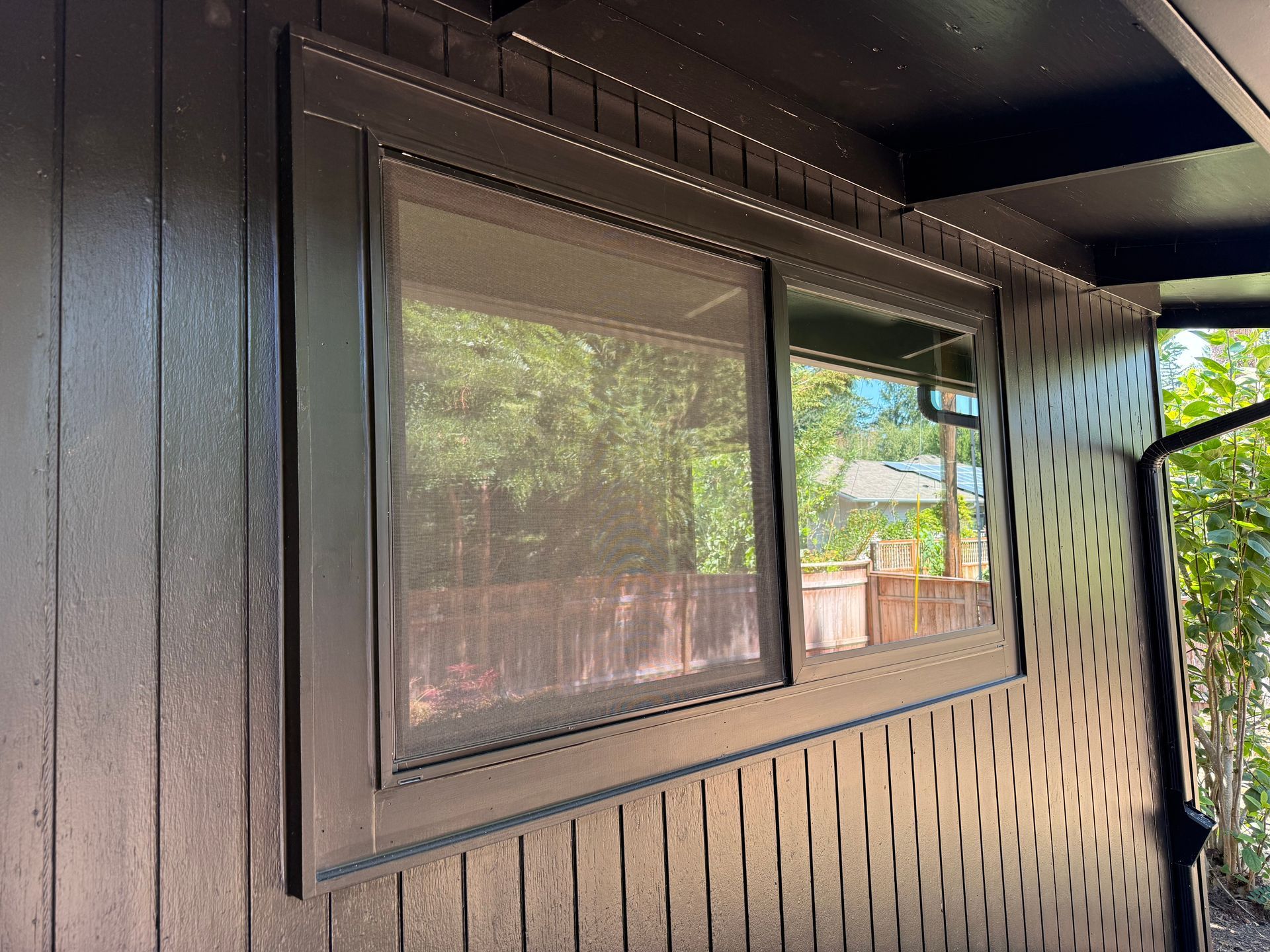 Black framed window with a screen, set in dark brown siding, with a view of trees and a fence.