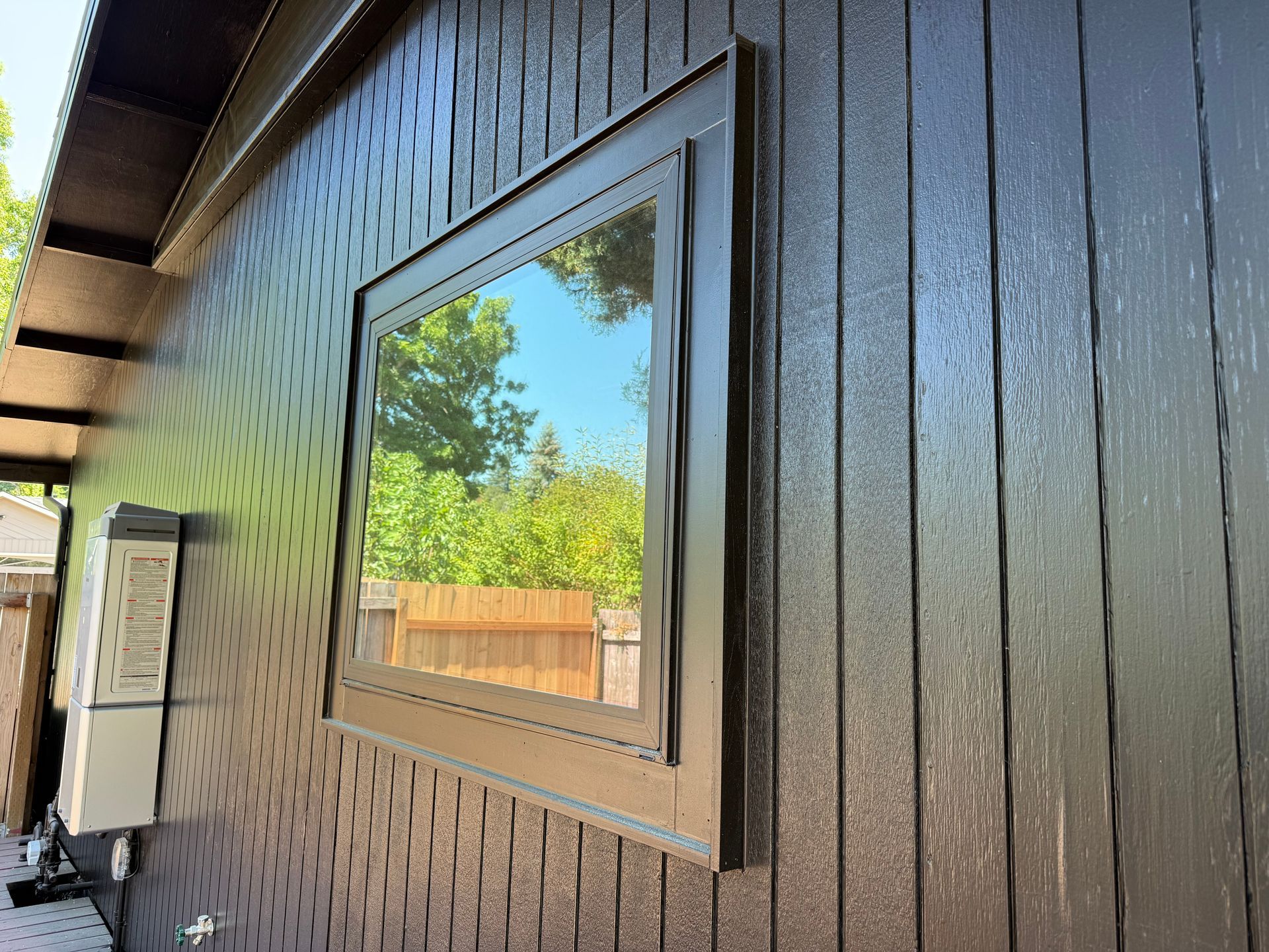 Brown-sided building with a rectangular window reflecting trees and a fence.