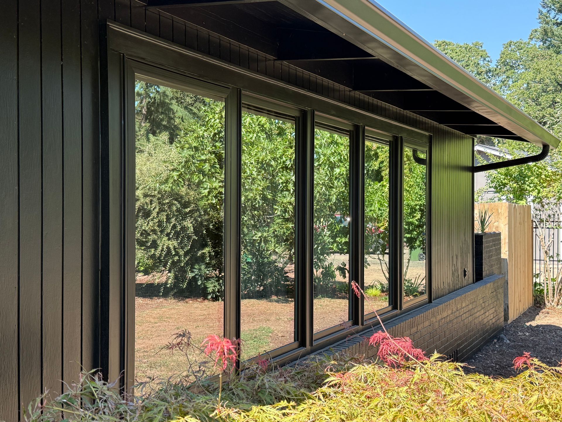 Black framed windows on a dark-sided building reflect a lush green yard; a black roof overhangs the windows.