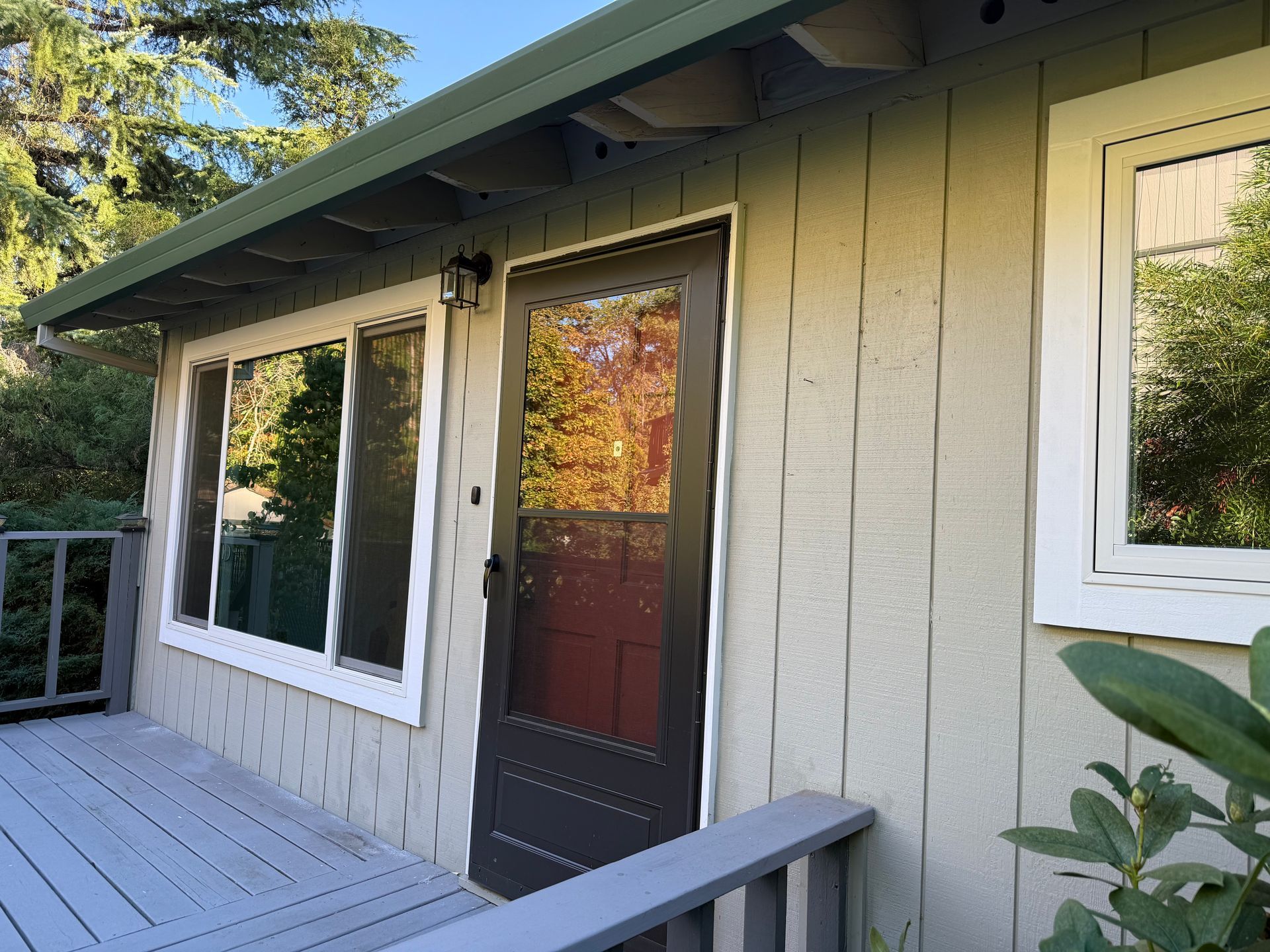 Exterior of a light green house with a dark door and white-framed windows, surrounded by a deck and trees.
