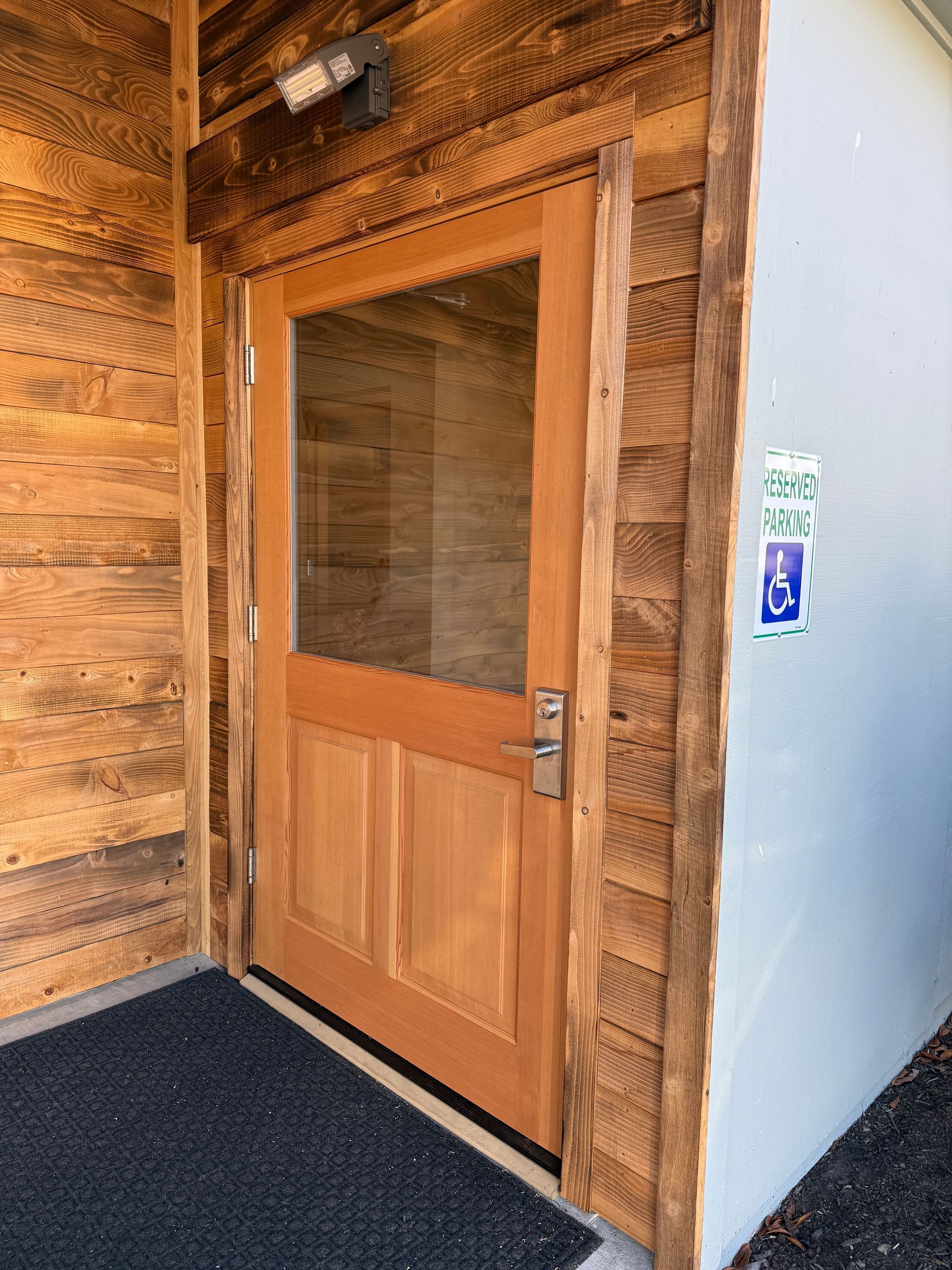Wooden door with glass panel, recessed in a wood and light-colored wall, with a door mat.