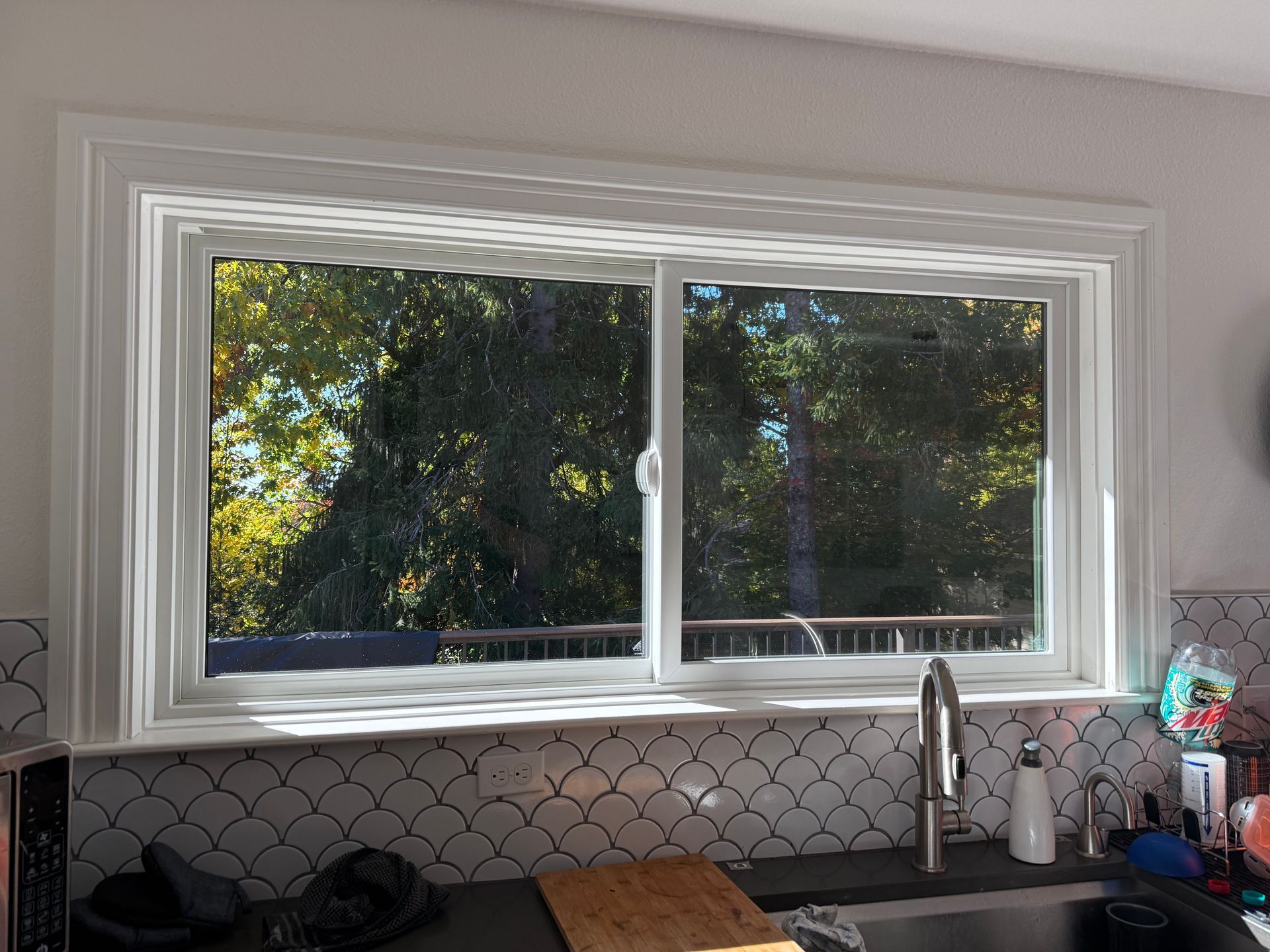 White-framed kitchen window with view of trees and a deck. Sunlight streams in.