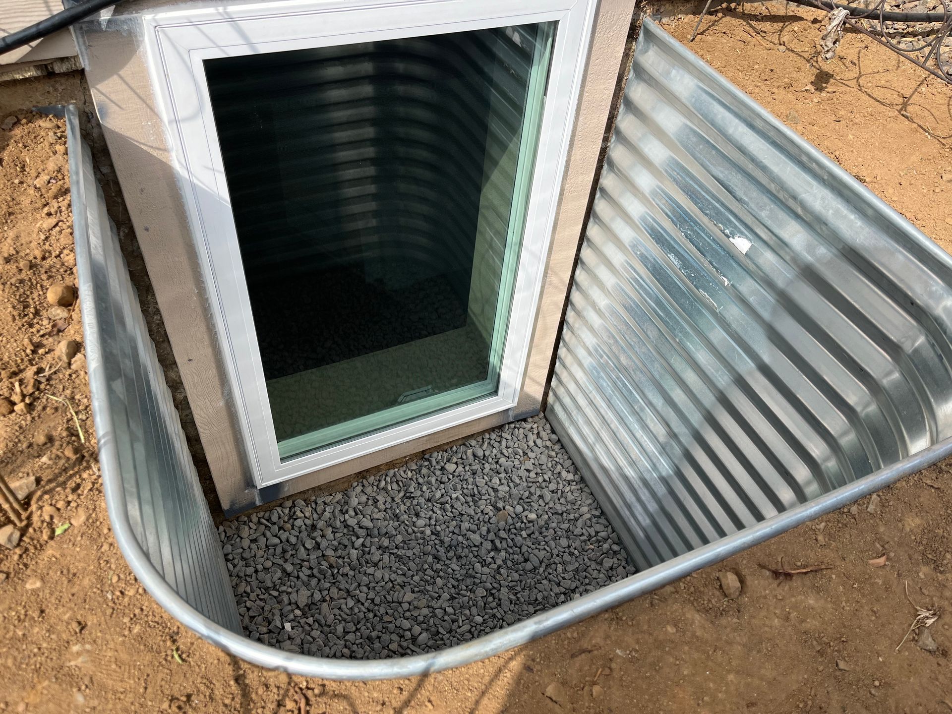A corrugated metal window well installed around a basement window, filled with gravel at the bottom.
