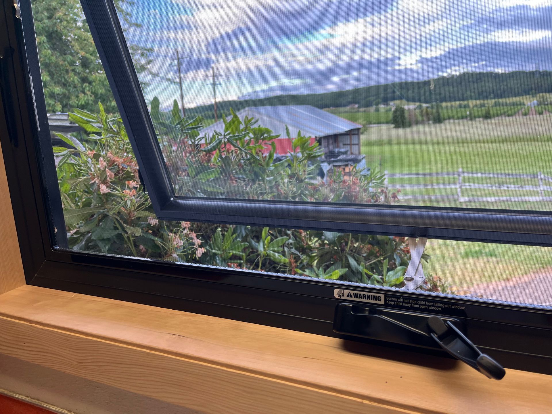 Open window with screen, black frame, overlooking a green landscape and a distant house.