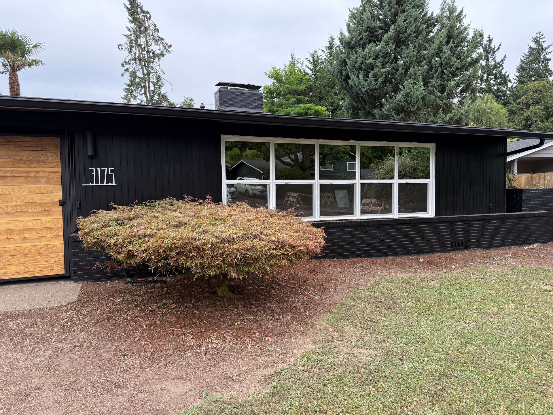 Black house with light wood door, white-framed windows, and a small bush in front.