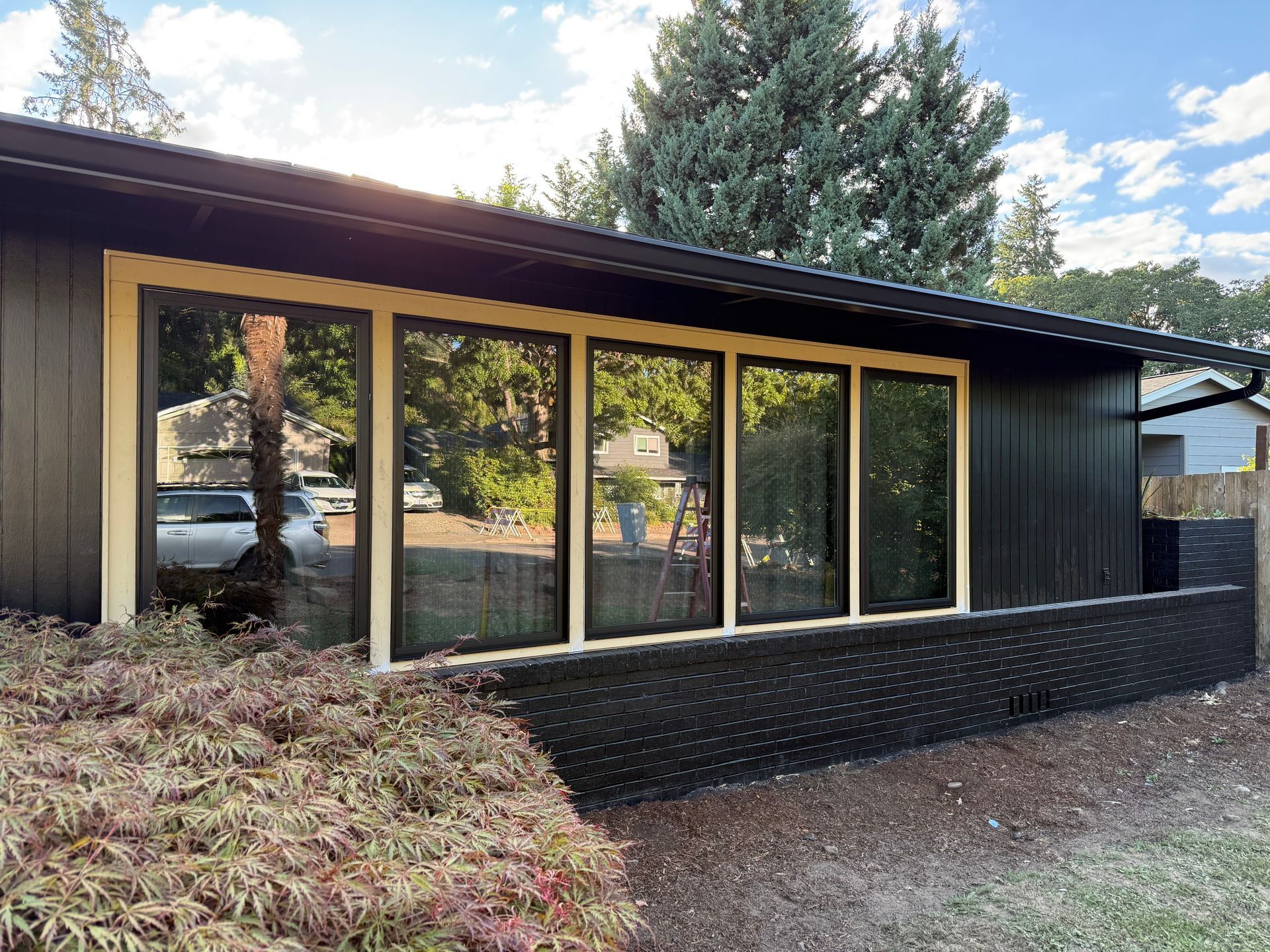 Black house exterior with large windows reflecting surroundings, tan trim, and a low black brick wall.
