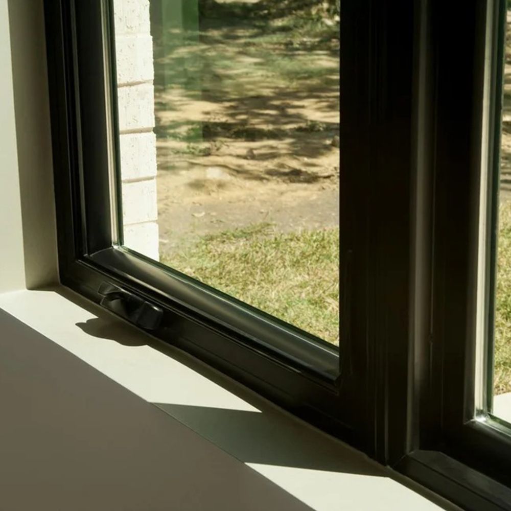 Close-up of a black window frame, with a handle, overlooking a grassy yard under sunlight.