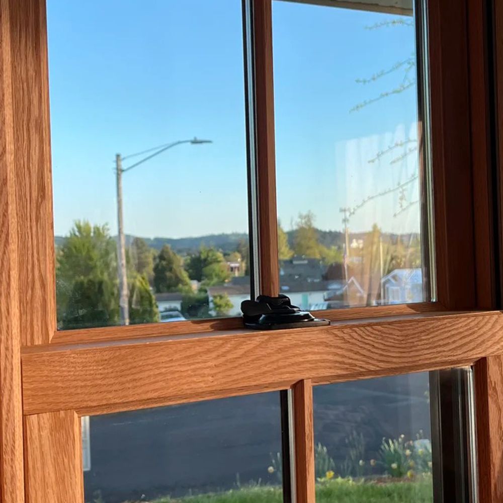 View through a wooden window: street, houses, trees, and blue sky on a sunny day.