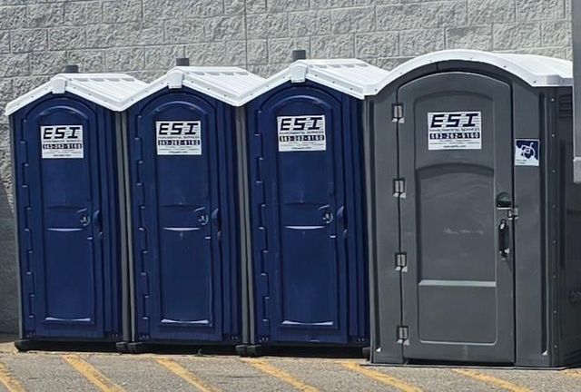 Four portable toilets, three blue and one gray, against a white wall.