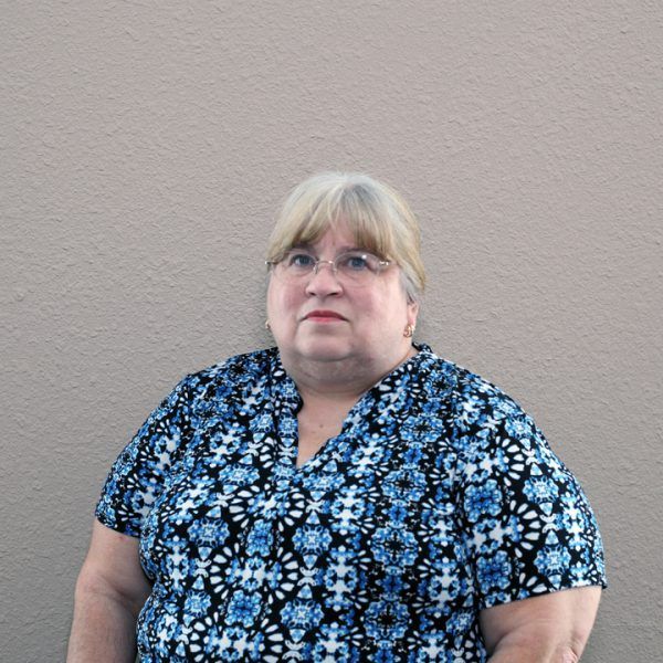 A woman wearing glasses and a blue shirt is standing in front of a wall.