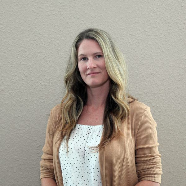 A woman wearing a brown cardigan and a white shirt is standing in front of a wall.