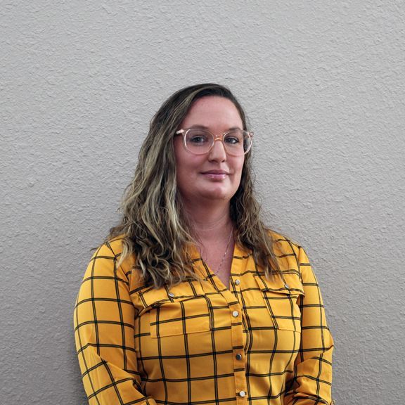 A woman wearing glasses and a yellow plaid shirt is standing in front of a wall.