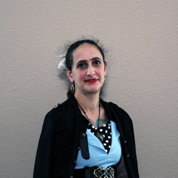 A woman wearing a black jacket and a blue shirt is standing in front of a wall