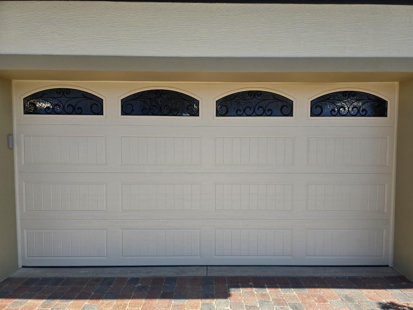 A white garage door with arched windows is sitting on a brick driveway.