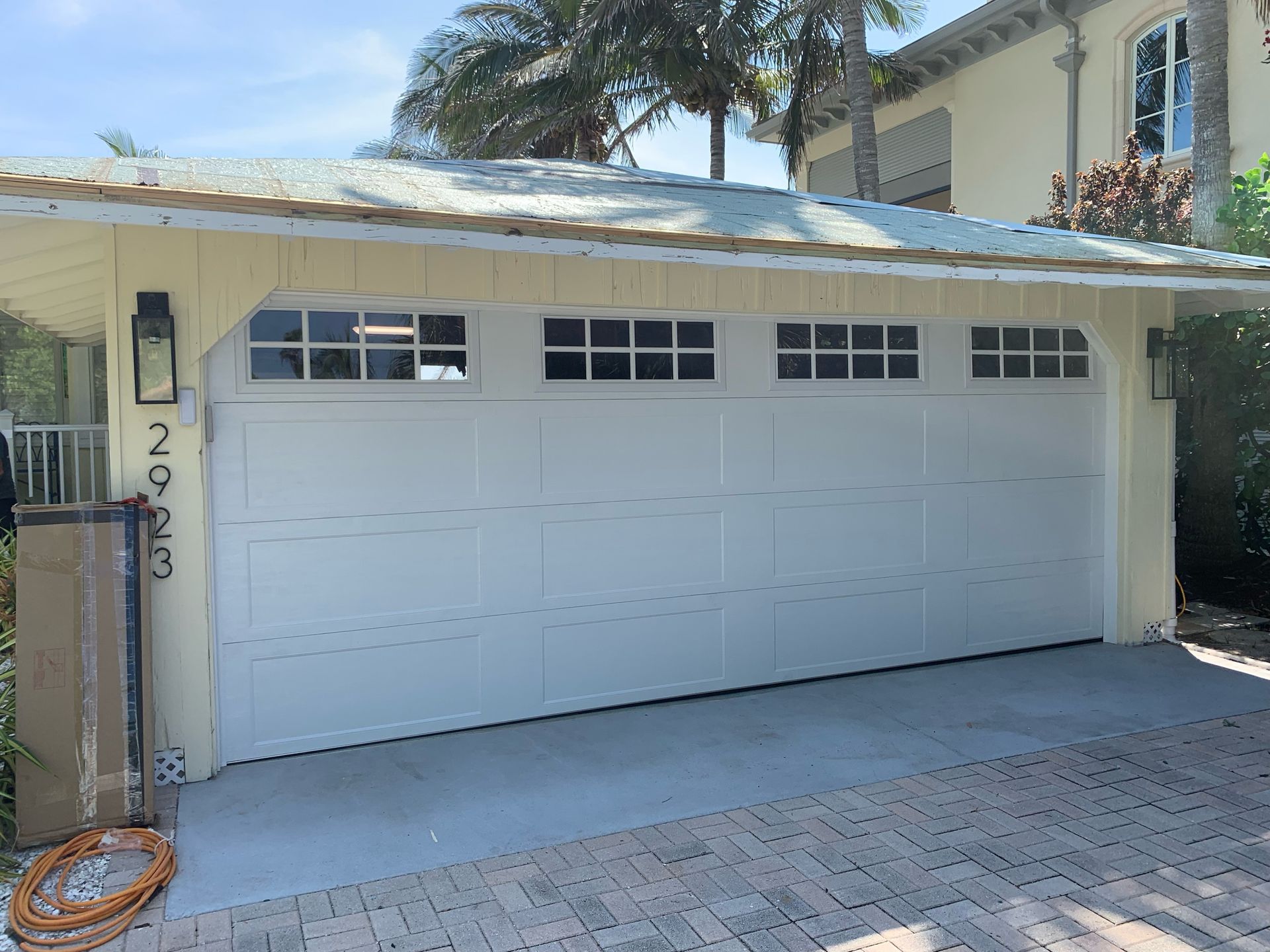 White two-car garage with a closed door, driveway, and palm trees outside a house.