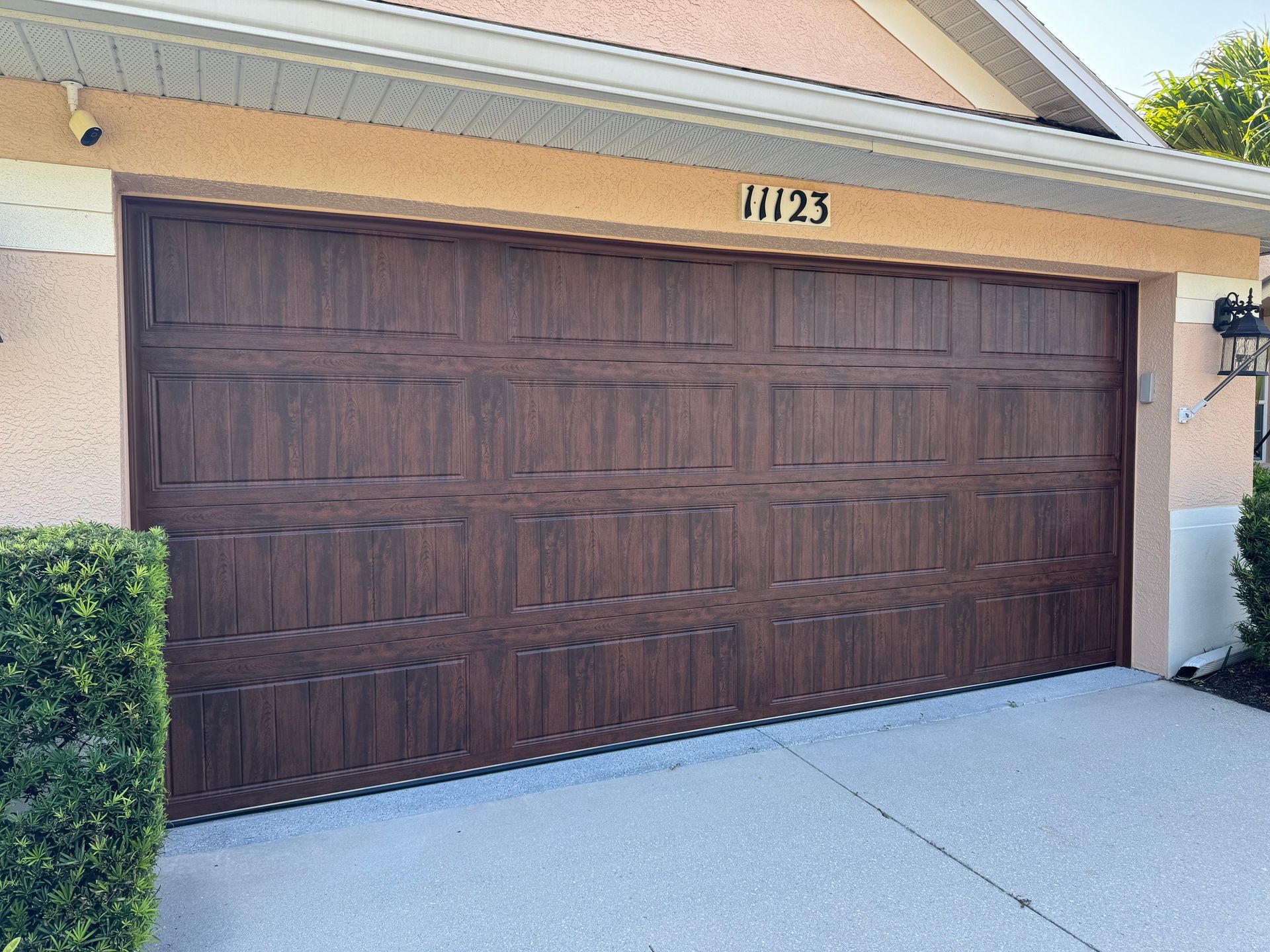 Dark brown double garage door with house number 1123 above it, beside a driveway and trimmed hedge.