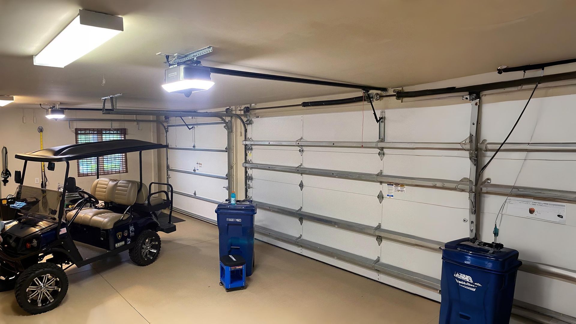 Wide garage with a parked black golf cart, blue trash bins, and a closed segmented garage door.