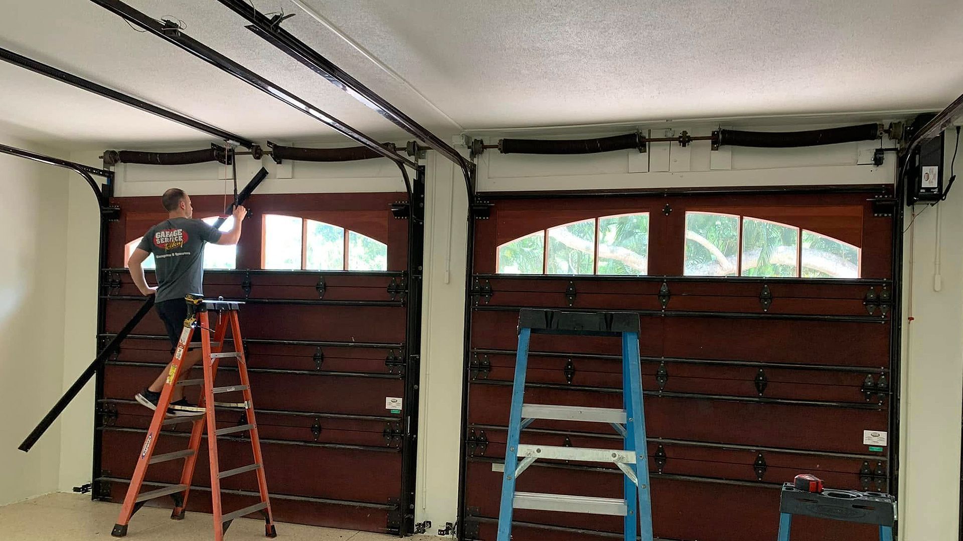 Two ladders in a garage with overhead door tracks and windows; a person stands on the left ladder near the ceiling.