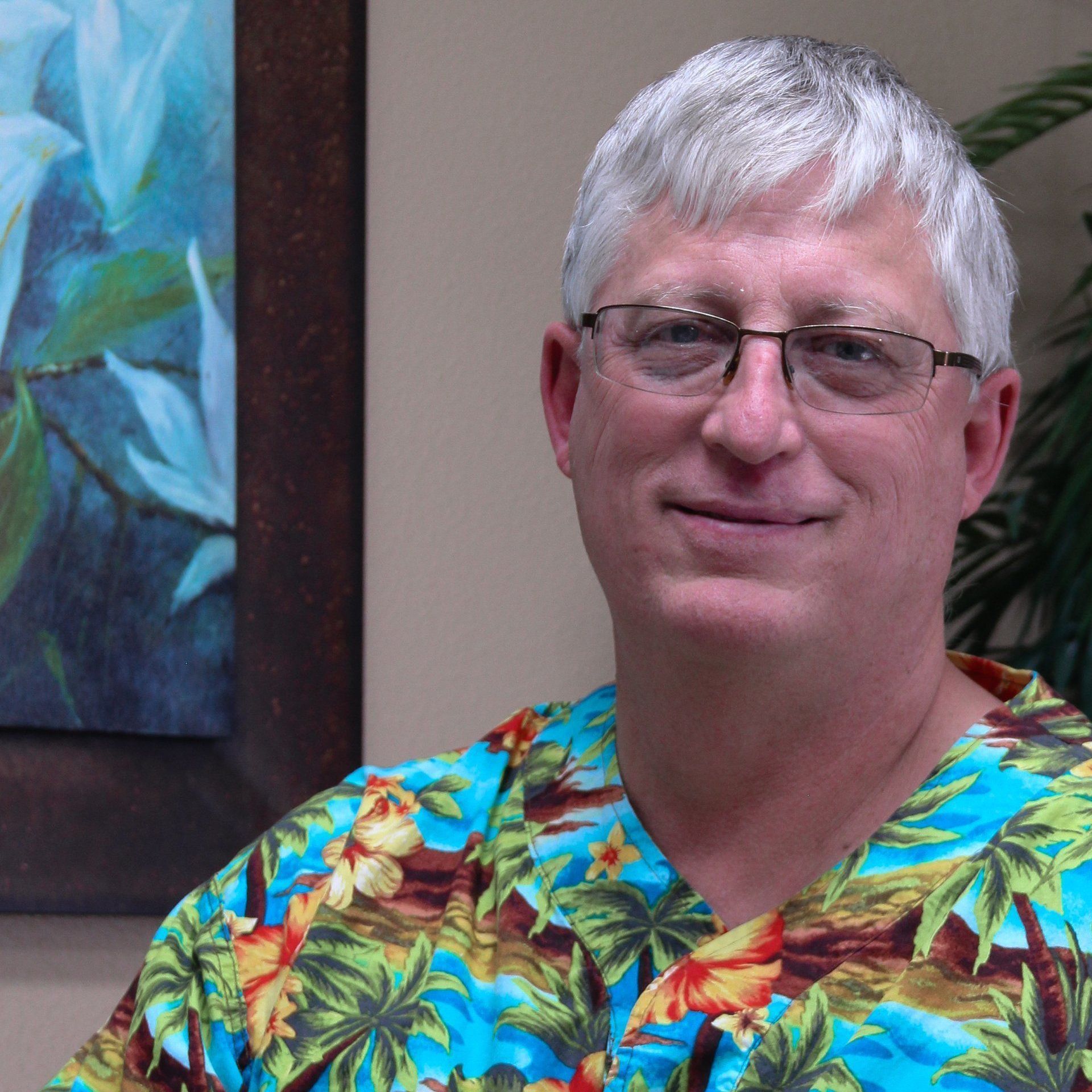 A man wearing scrubs and glasses is smiling in front of a painting