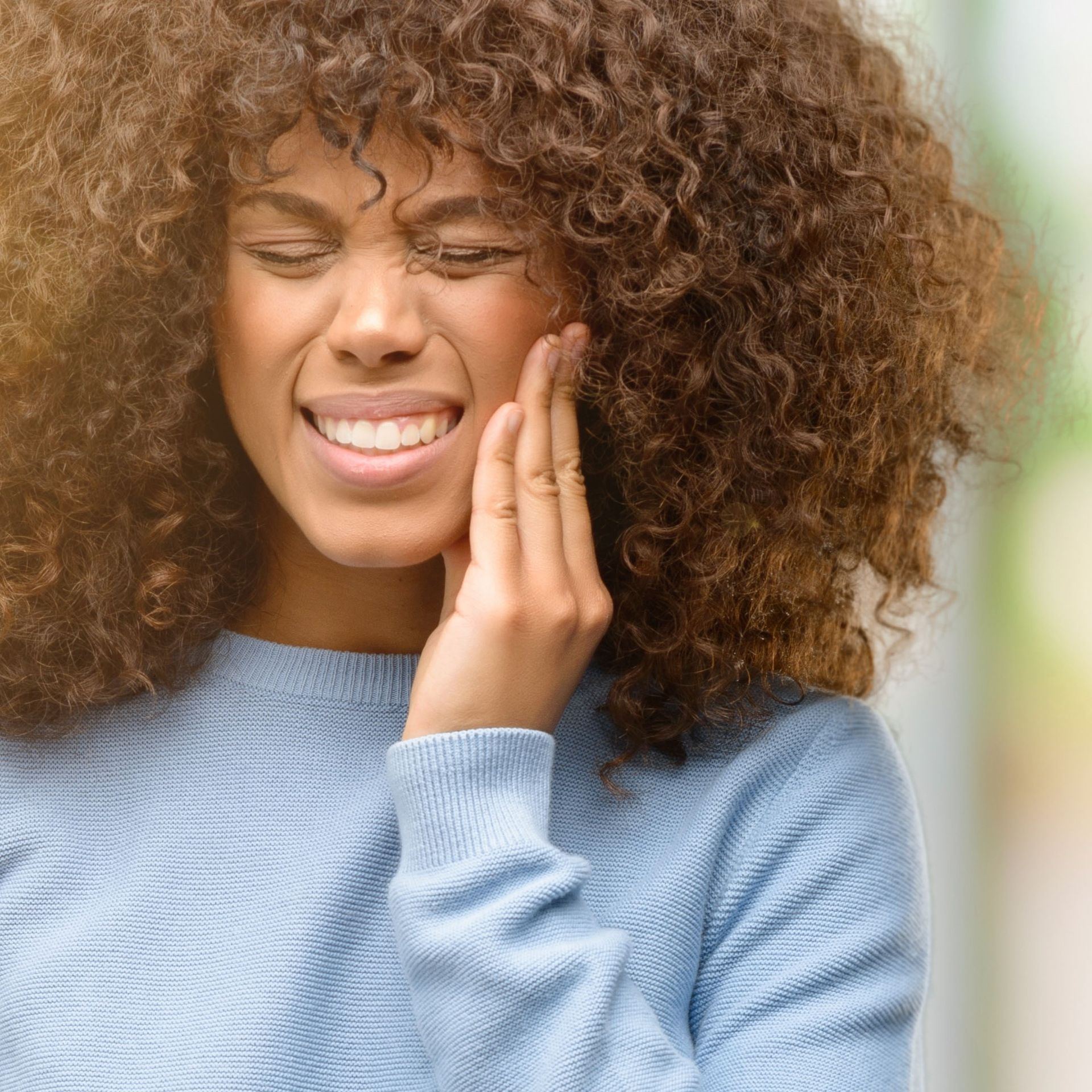 A woman with curly hair is smiling and touching her face.