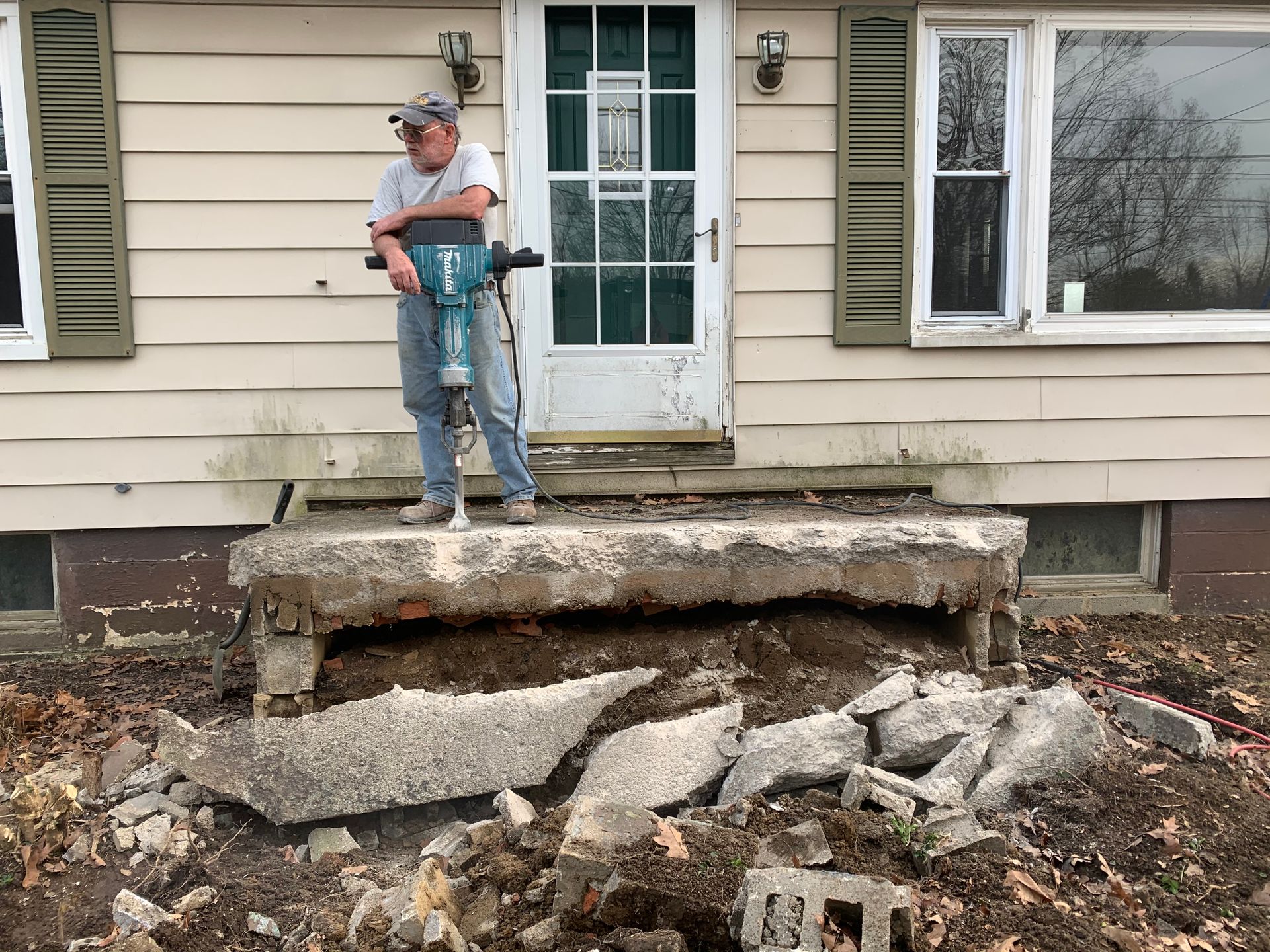 Man using jackhammer to demolish a concrete porch in front of a house.